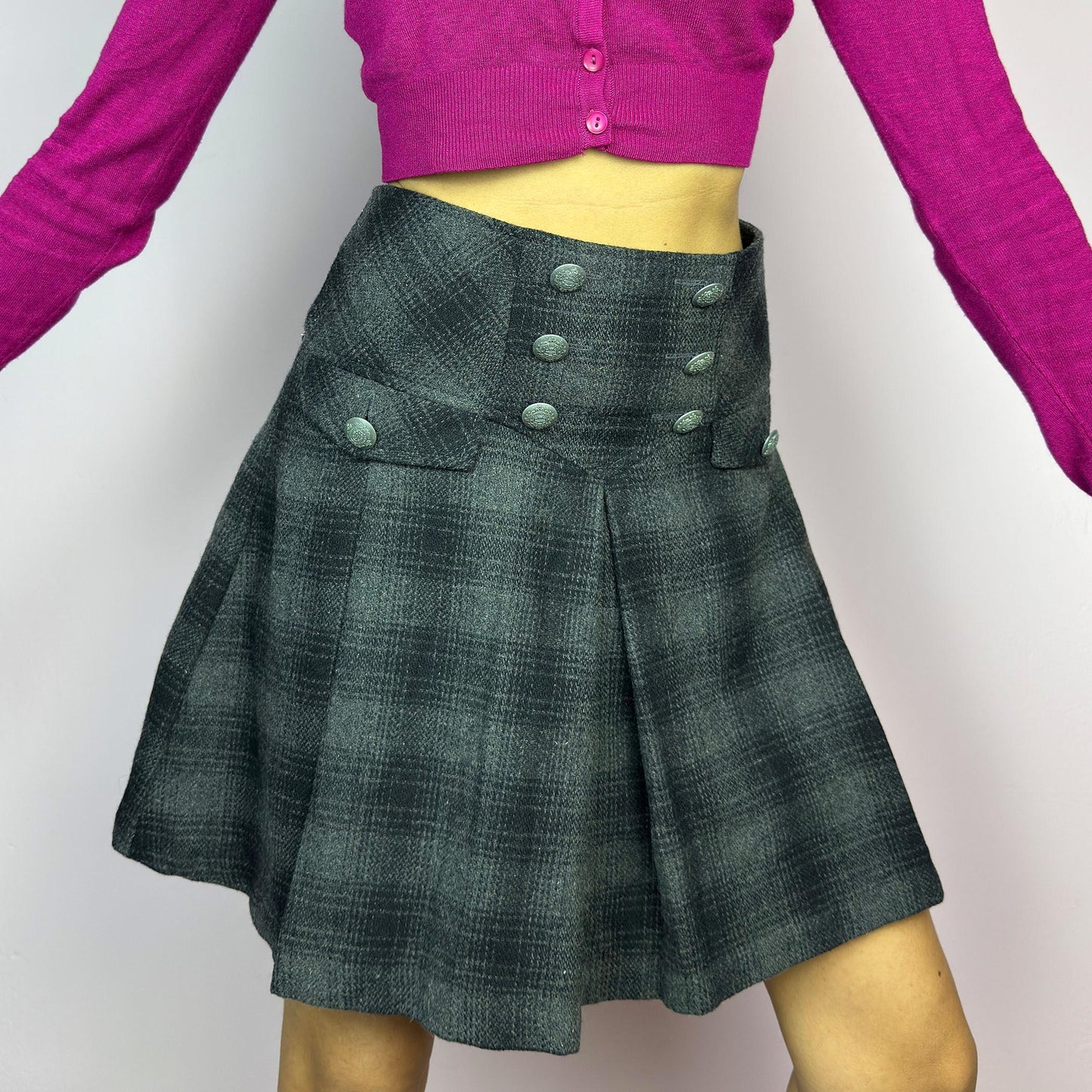 The Y2K Gray Plaid Pleated Mini Skirt is a vintage 2000s essential, featuring a structured pleated design with decorative button details. Made from a thick woven fabric with a black plaid pattern, it is ideal for winter outfits. adult world shop