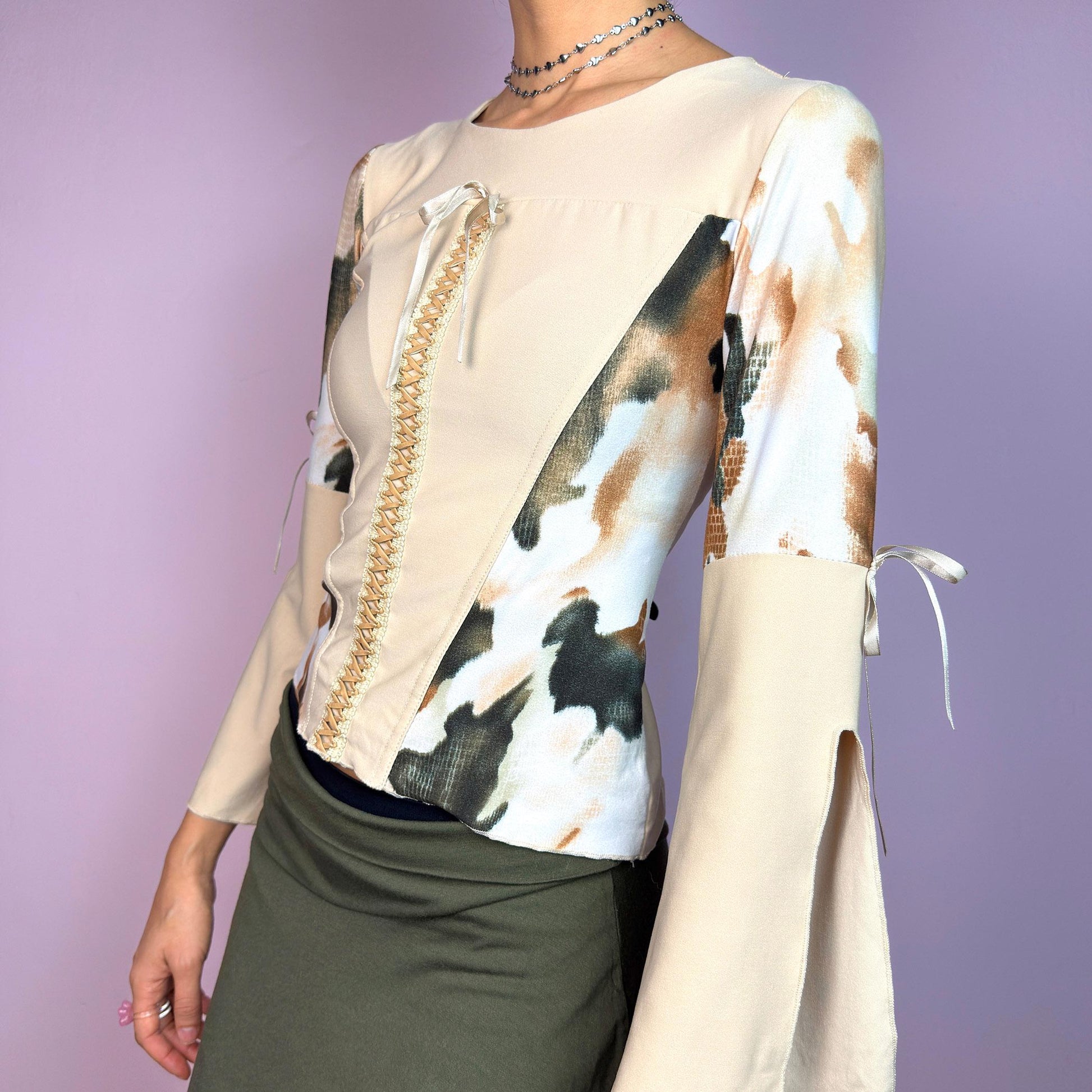 The Y2K Beige Bell Sleeve Top is a vintage 2000s blouse crafted from a stretchy fabric and featuring an abstract print on the sleeves and side panels, contrasted with a solid beige bodice. The design includes a lace-up front detail. adult world shop