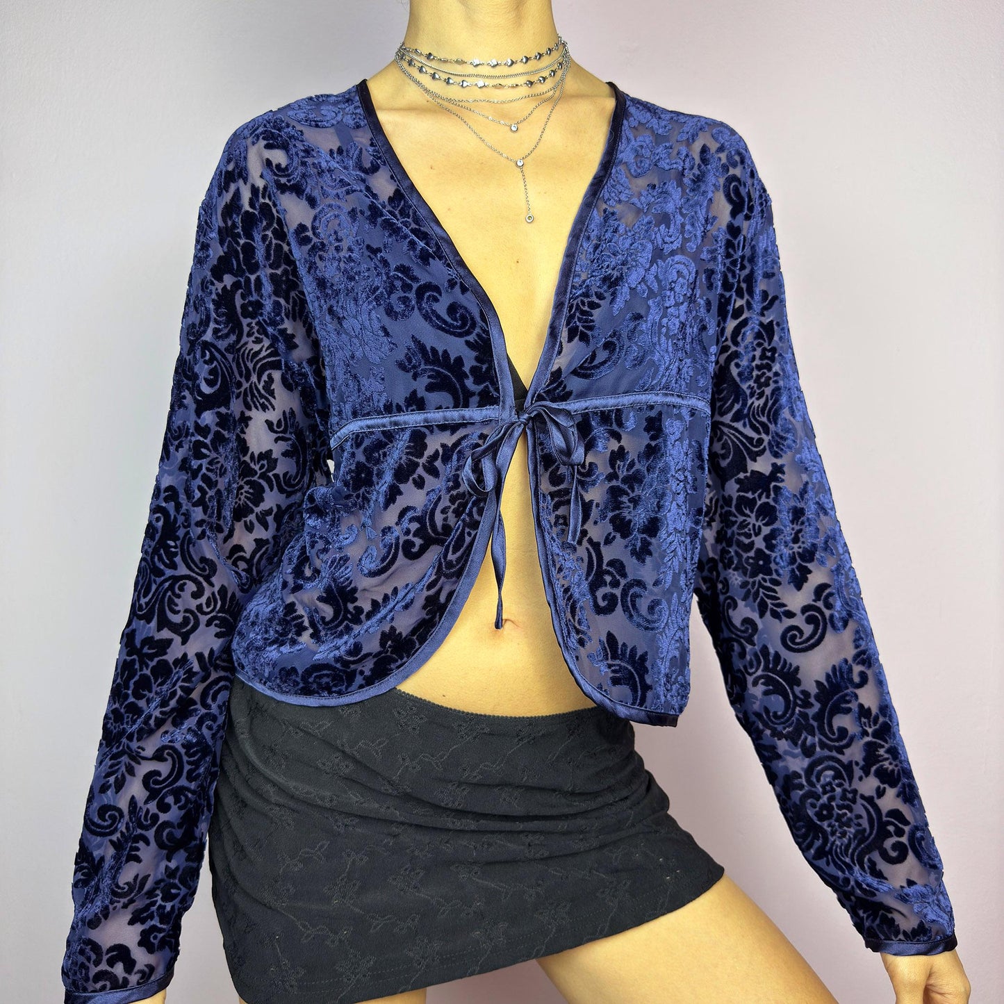 The Vintage 90s Blue Velvet Sheer Blouse is a romantic sheer velvet top featuring a flocked baroque floral pattern on deep navy blue. Designed with a plunging V-neckline, long sleeves, and a delicate tie-front closure, this blouse is perfect for night-out looks. adult world shop