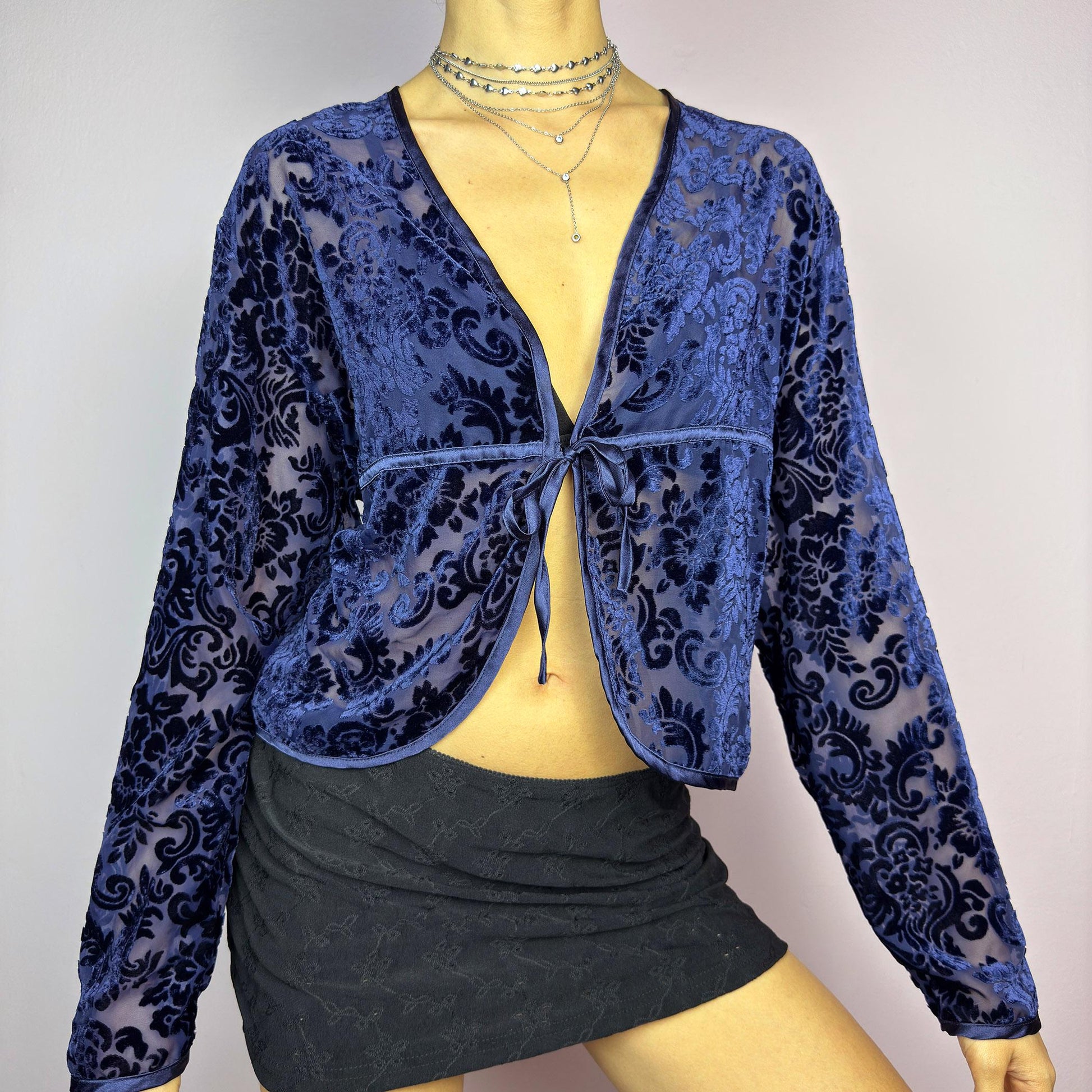 The Vintage 90s Blue Velvet Sheer Blouse is a romantic sheer velvet top featuring a flocked baroque floral pattern on deep navy blue. Designed with a plunging V-neckline, long sleeves, and a delicate tie-front closure, this blouse is perfect for night-out looks. adult world shop