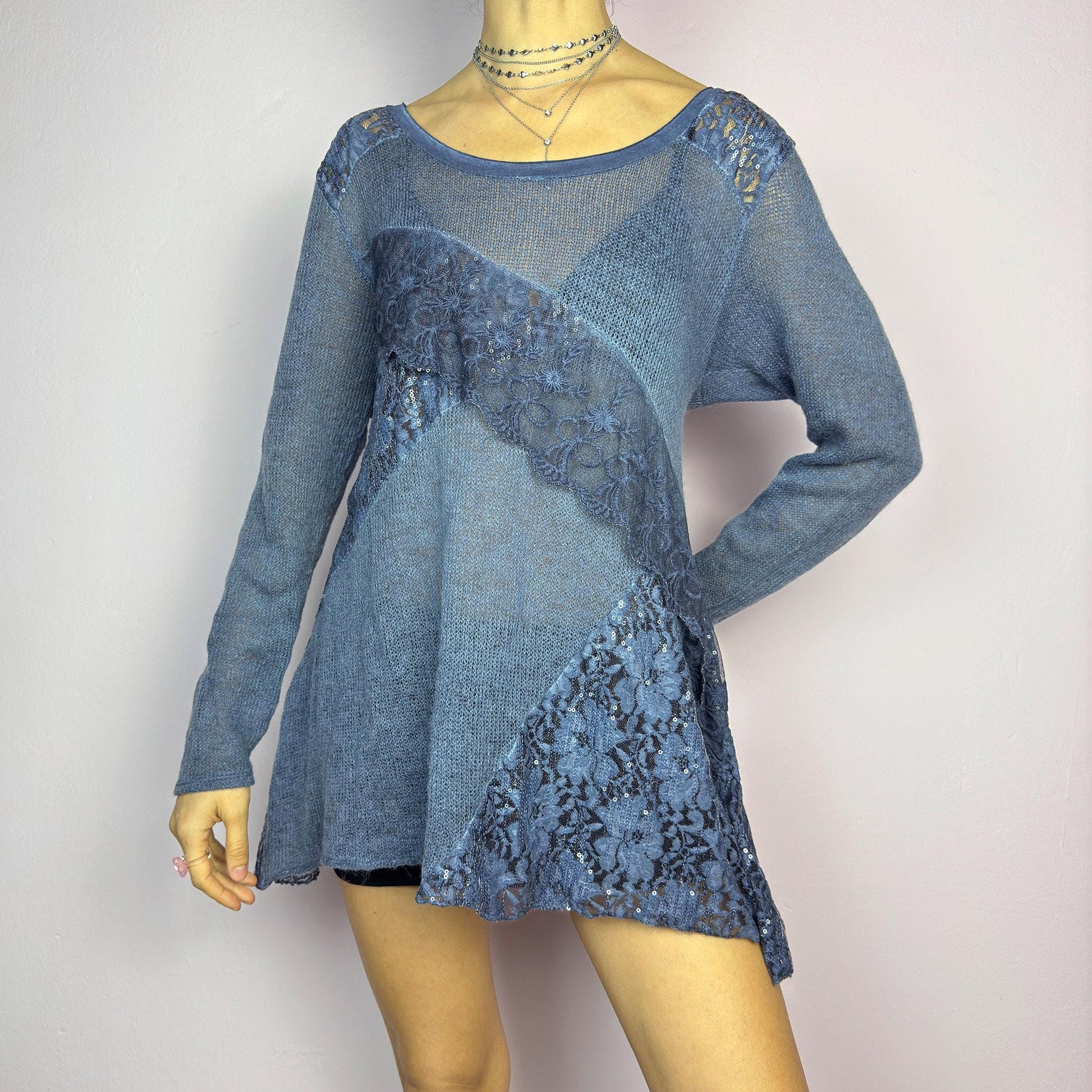 The Y2K Blue Lace Asymmetric Sweater is a vintage 2000s piece featuring a semi-sheer knit design with delicate lace and sequin paneling in an asymmetrical pattern. Designed with a scoop neckline, long sleeves, and a flowy silhouette, this sweater tunic adds a dreamy, romantic touch to any outfit. Perfect for layering in autumn and winter. adult world shop