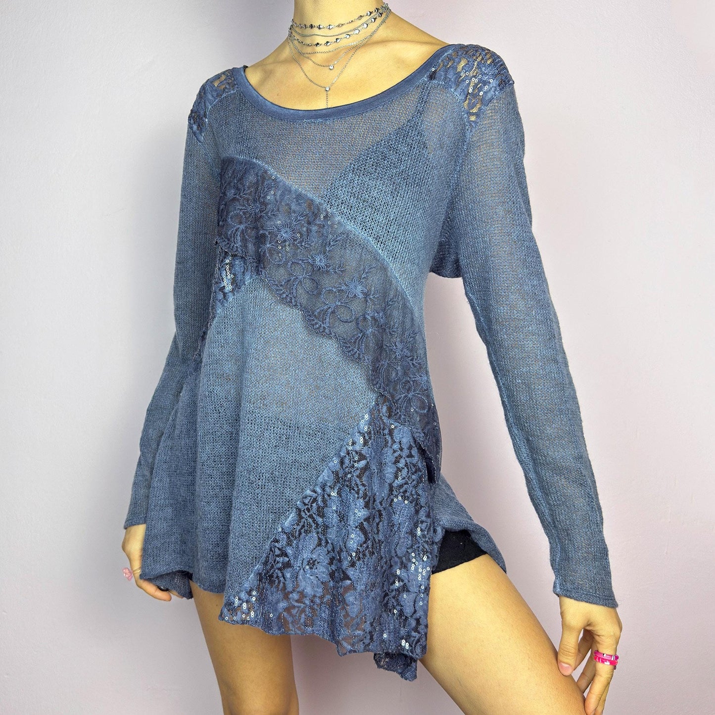 The Y2K Blue Lace Asymmetric Sweater is a vintage 2000s piece featuring a semi-sheer knit design with delicate lace and sequin paneling in an asymmetrical pattern. Designed with a scoop neckline, long sleeves, and a flowy silhouette, this sweater tunic adds a dreamy, romantic touch to any outfit. Perfect for layering in autumn and winter. adult world shop