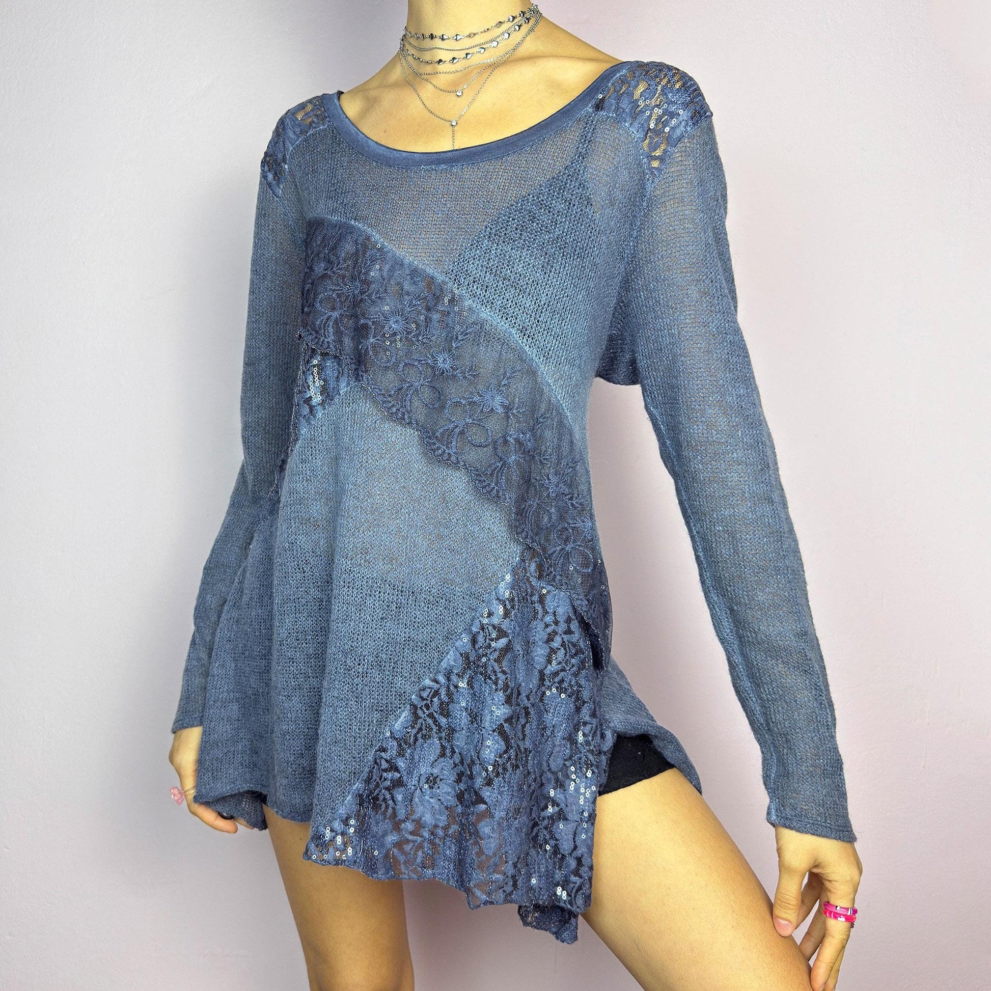 The Y2K Blue Lace Asymmetric Sweater is a vintage 2000s piece featuring a semi-sheer knit design with delicate lace and sequin paneling in an asymmetrical pattern. Designed with a scoop neckline, long sleeves, and a flowy silhouette, this sweater tunic adds a dreamy, romantic touch to any outfit. Perfect for layering in autumn and winter. adult world shop