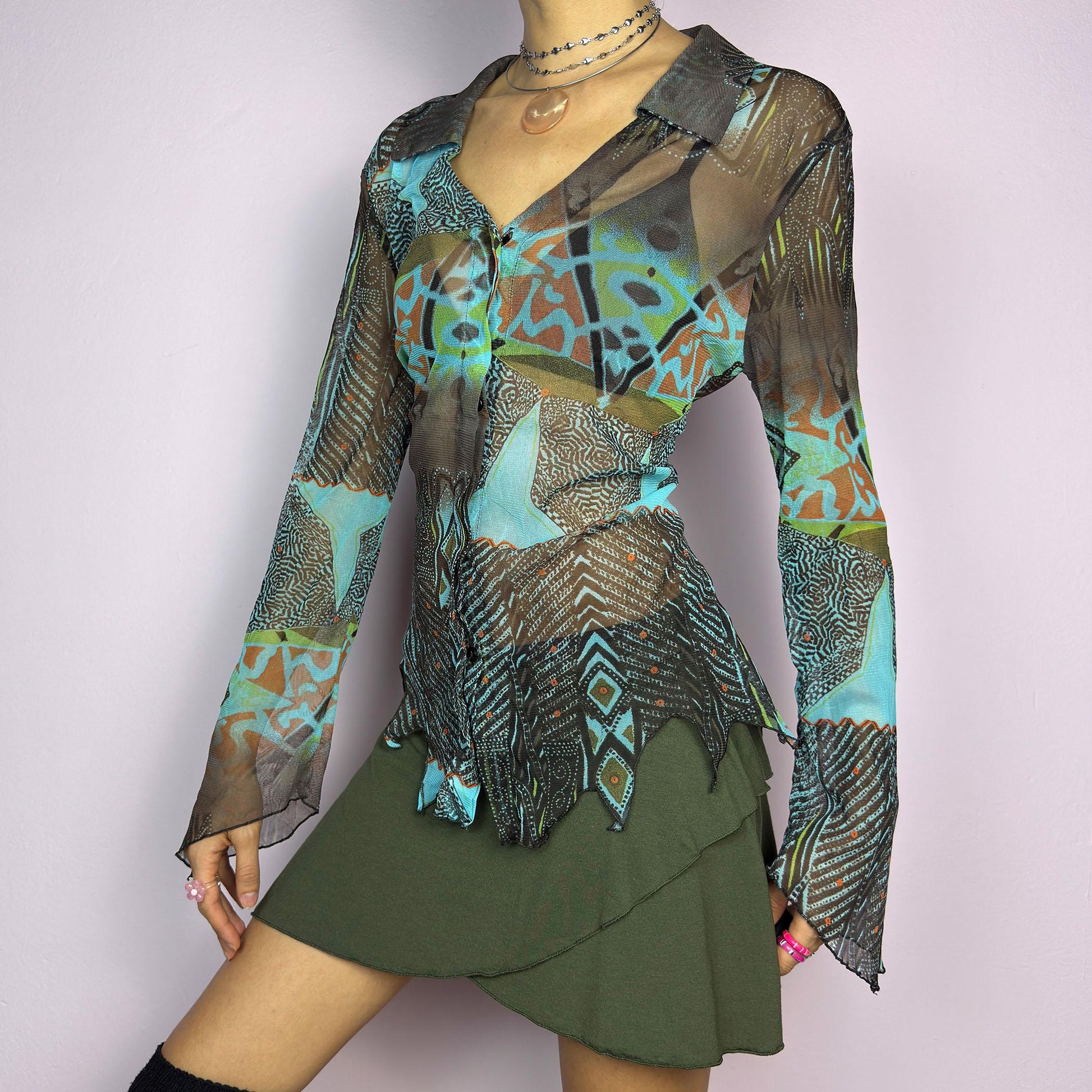 The Y2K Abstract Mesh Blouse is a vintage 2000s edgy and whimsical piece featuring a semi-sheer mesh fabric with bold, abstract patterns in earthy and blue tones and a button-front closure. The long flared sleeves. adult world shop