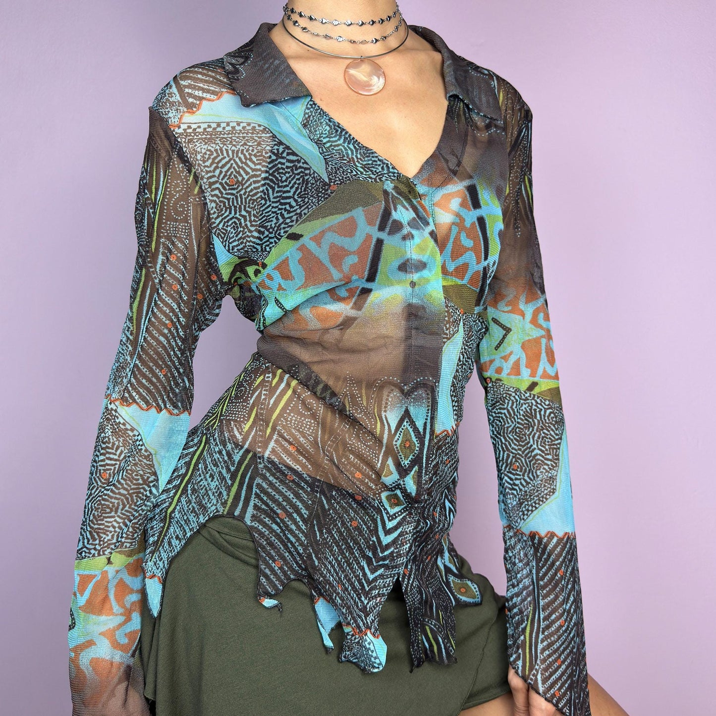 The Y2K Abstract Mesh Blouse is a vintage 2000s edgy and whimsical piece featuring a semi-sheer mesh fabric with bold, abstract patterns in earthy and blue tones and a button-front closure. The long flared sleeves. adult world shop