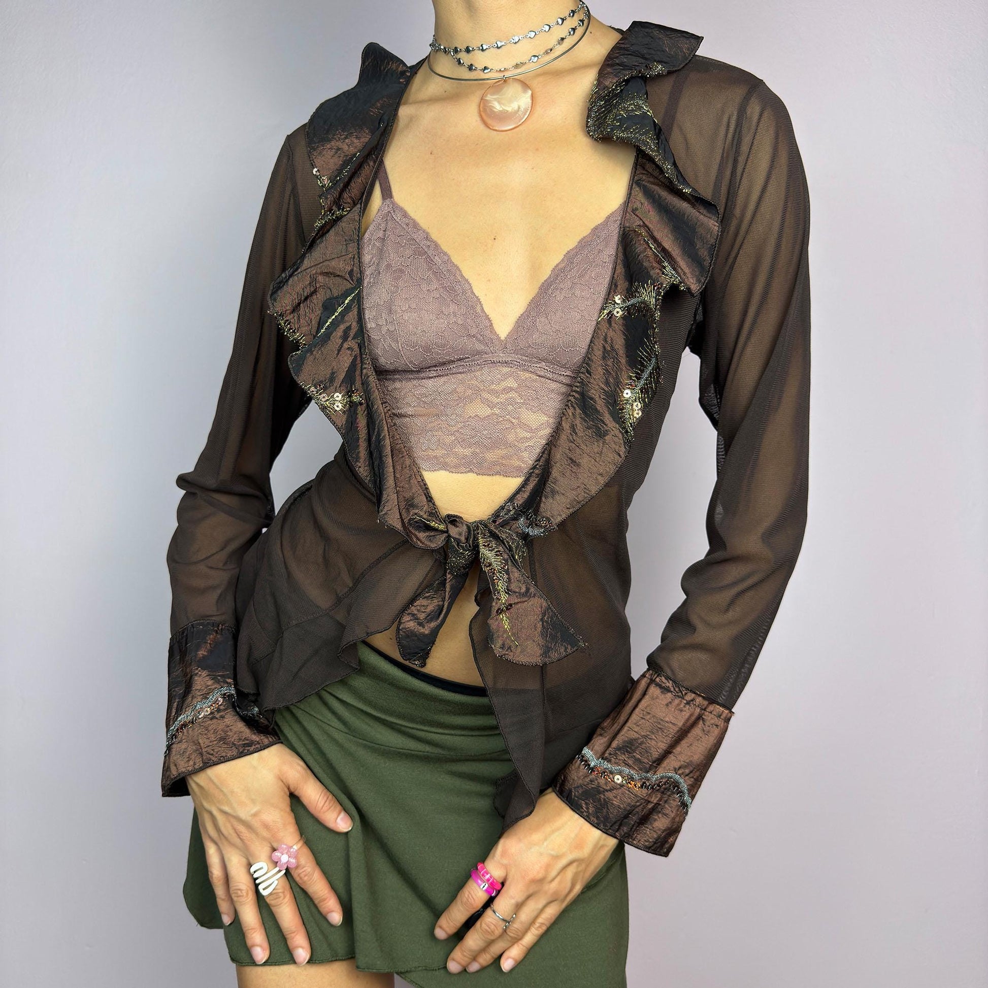 The Y2K Brown Mesh Tie Front Top is a vintage 2000s elegant and flirty piece. This semi-sheer mesh blouse features a tie-front closure and a cascade of ruffle detailing along the neckline and cuffs. adult world shop