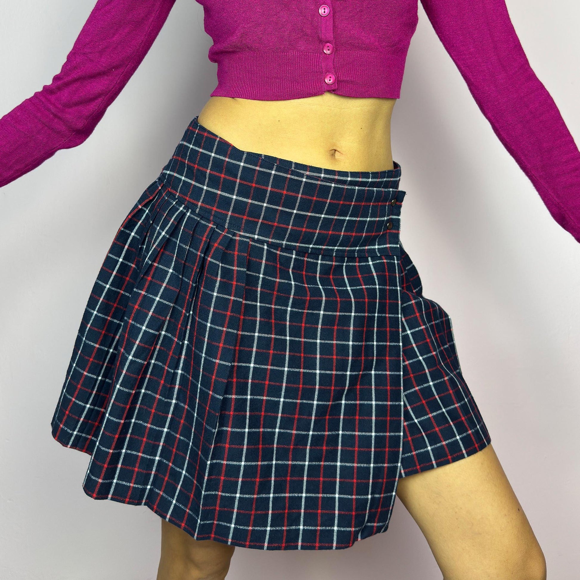 The Y2K Plaid Pleated Wrap Mini Skirt is a vintage 2000s piece featuring a wrap-style waistband with snap button details. Made from a plaid fabric in navy blue tones, it has structured pleats that create a classic silhouette. adult world shop