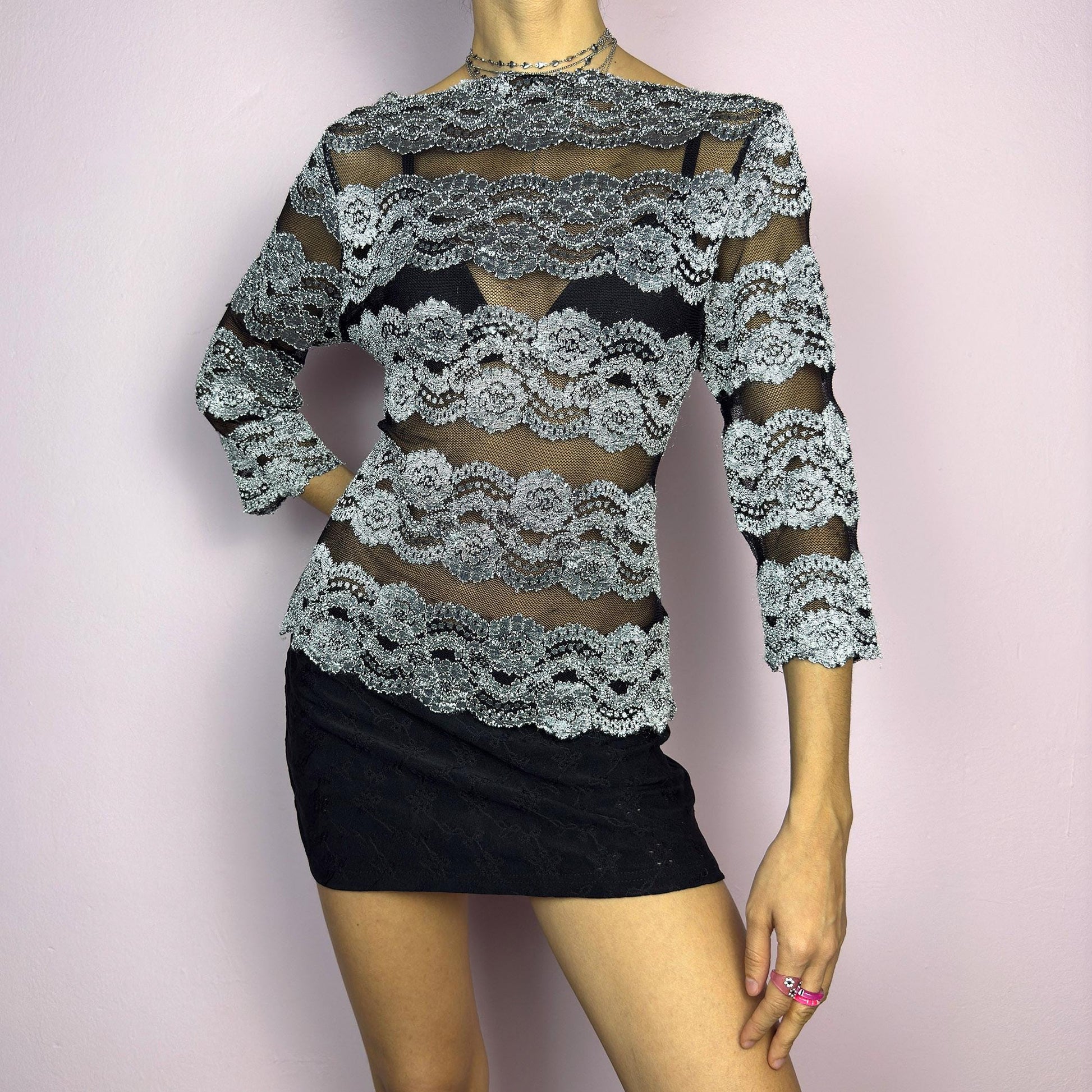 The Y2K Lace Mesh Black Top is a vintage 2000s statement sheer blouse featuring a delicate black mesh base with intricate silver lace embroidery in horizontal stripes. Designed with a fitted silhouette and three-quarter sleeves, this top is perfect for a romantic night or a bold evening outfit.
