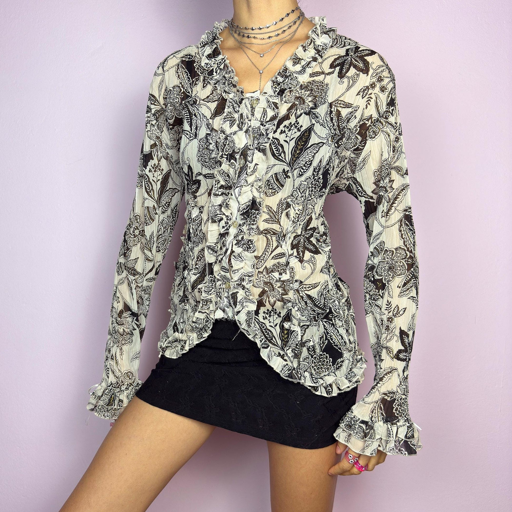 The Y2K Cream Ruffle Blouse is a vintage 2000s sheer top featuring an intricate black and white floral print with cascading ruffle details along the front. Designed with a romantic and feminine touch, it has subtle glitter accents and delicate beaded details, a button-up front, flared sleeves with tie details, and a lightweight, slightly crinkled fabric. Perfect for romantic nights and casual-chic outfits. adult world shop