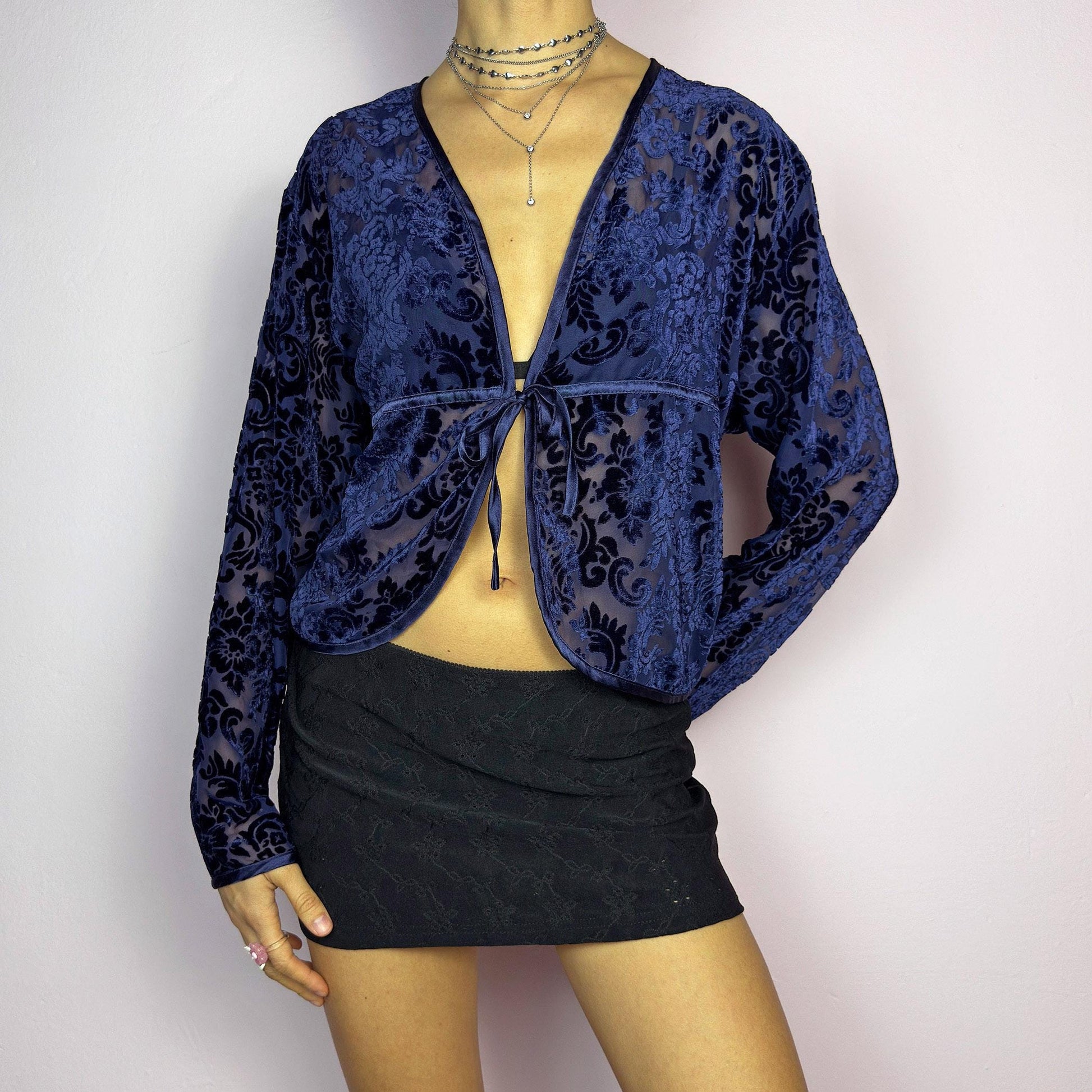 The Vintage 90s Blue Velvet Sheer Blouse is a romantic sheer velvet top featuring a flocked baroque floral pattern on deep navy blue. Designed with a plunging V-neckline, long sleeves, and a delicate tie-front closure, this blouse is perfect for night-out looks. adult world shop