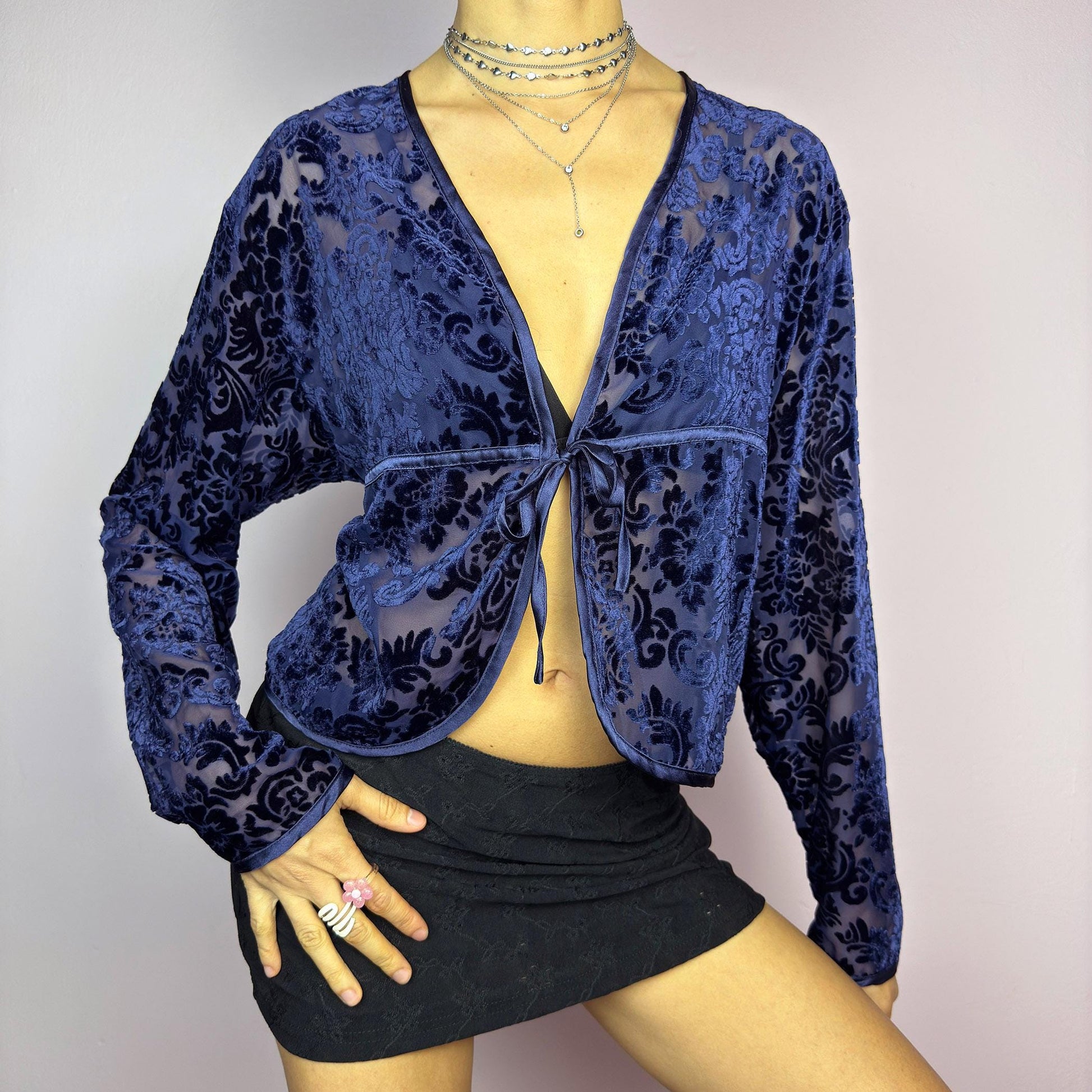 The Vintage 90s Blue Velvet Sheer Blouse is a romantic sheer velvet top featuring a flocked baroque floral pattern on deep navy blue. Designed with a plunging V-neckline, long sleeves, and a delicate tie-front closure, this blouse is perfect for night-out looks. adult world shop