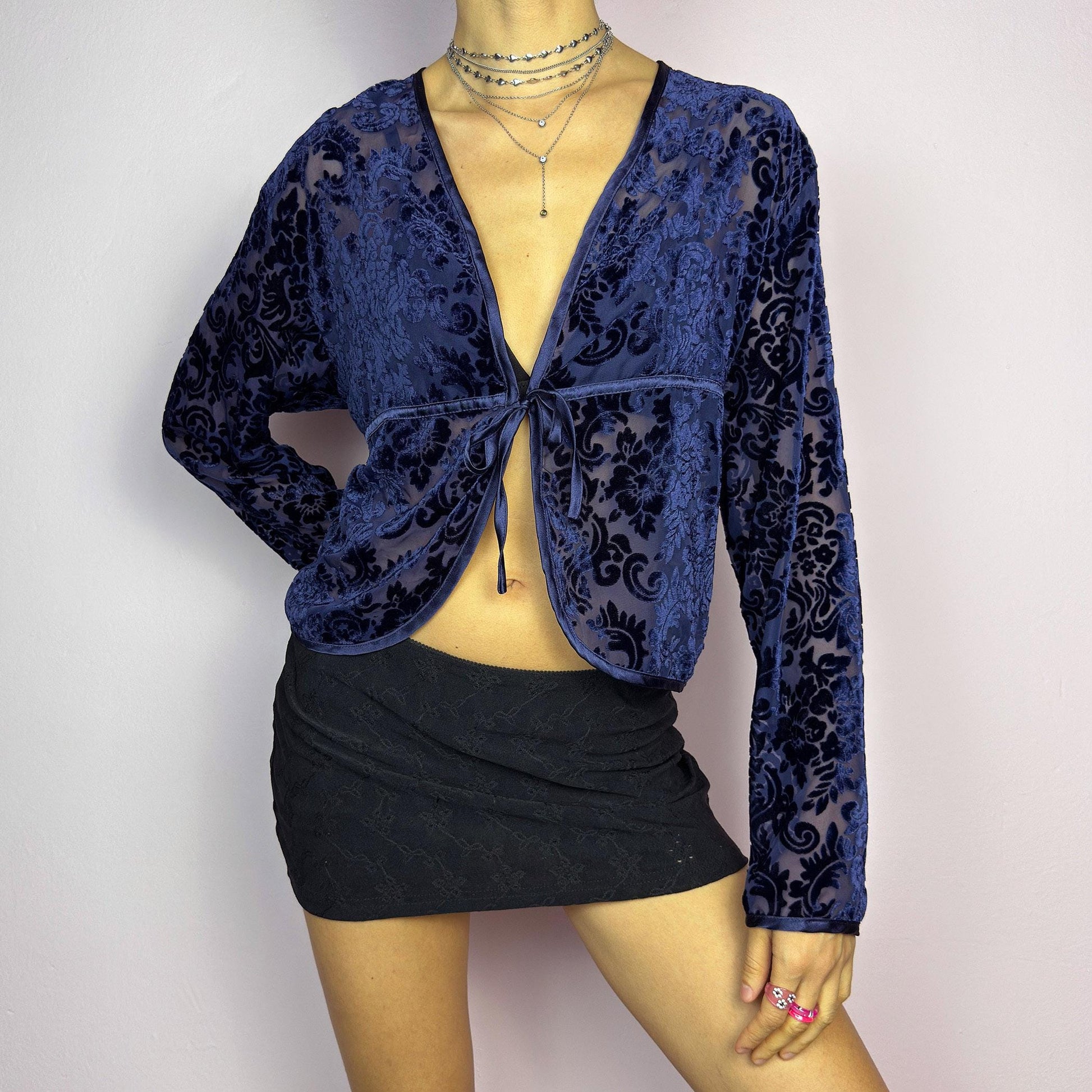 The Vintage 90s Blue Velvet Sheer Blouse is a romantic sheer velvet top featuring a flocked baroque floral pattern on deep navy blue. Designed with a plunging V-neckline, long sleeves, and a delicate tie-front closure, this blouse is perfect for night-out looks. adult world shop