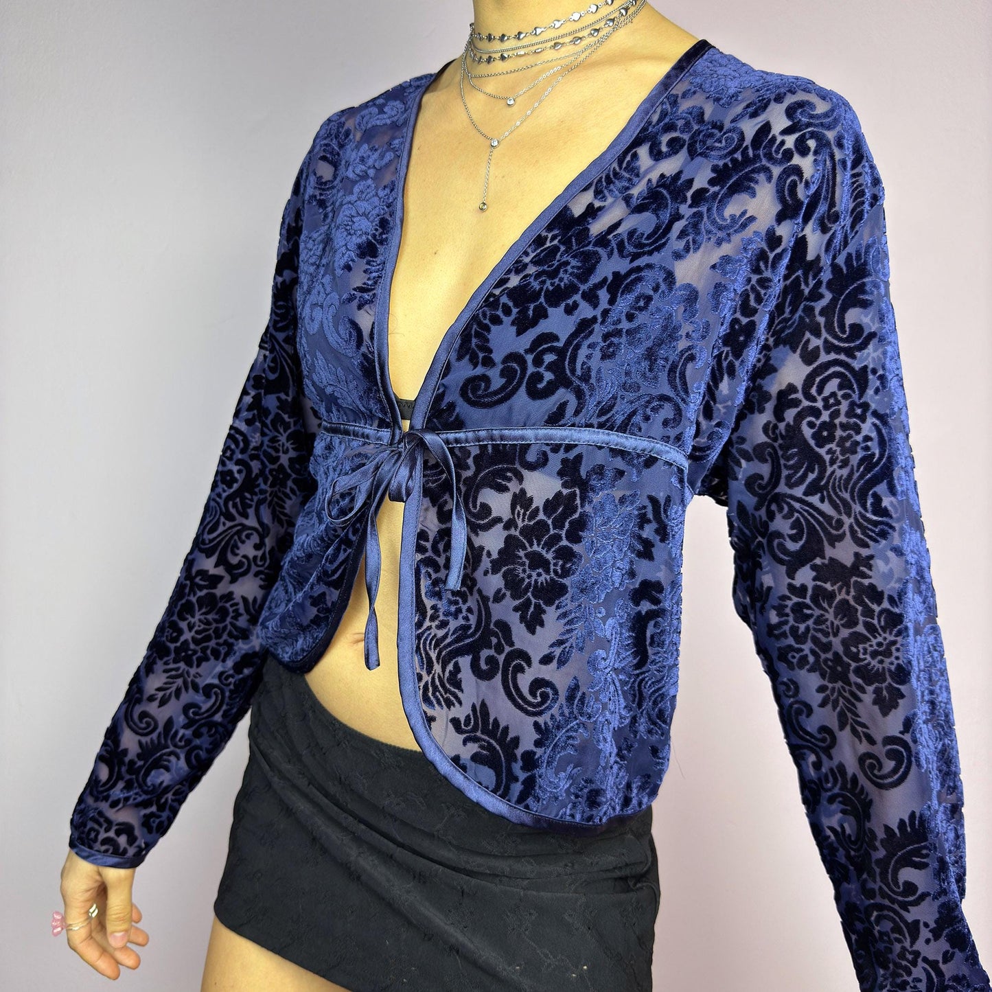 The Vintage 90s Blue Velvet Sheer Blouse is a romantic sheer velvet top featuring a flocked baroque floral pattern on deep navy blue. Designed with a plunging V-neckline, long sleeves, and a delicate tie-front closure, this blouse is perfect for night-out looks. adult world shop