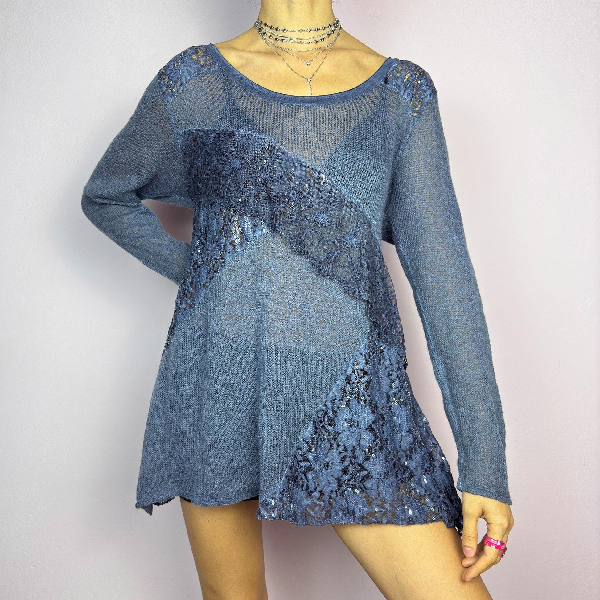 The Y2K Blue Lace Asymmetric Sweater is a vintage 2000s piece featuring a semi-sheer knit design with delicate lace and sequin paneling in an asymmetrical pattern. Designed with a scoop neckline, long sleeves, and a flowy silhouette, this sweater tunic adds a dreamy, romantic touch to any outfit. Perfect for layering in autumn and winter. adult world shop