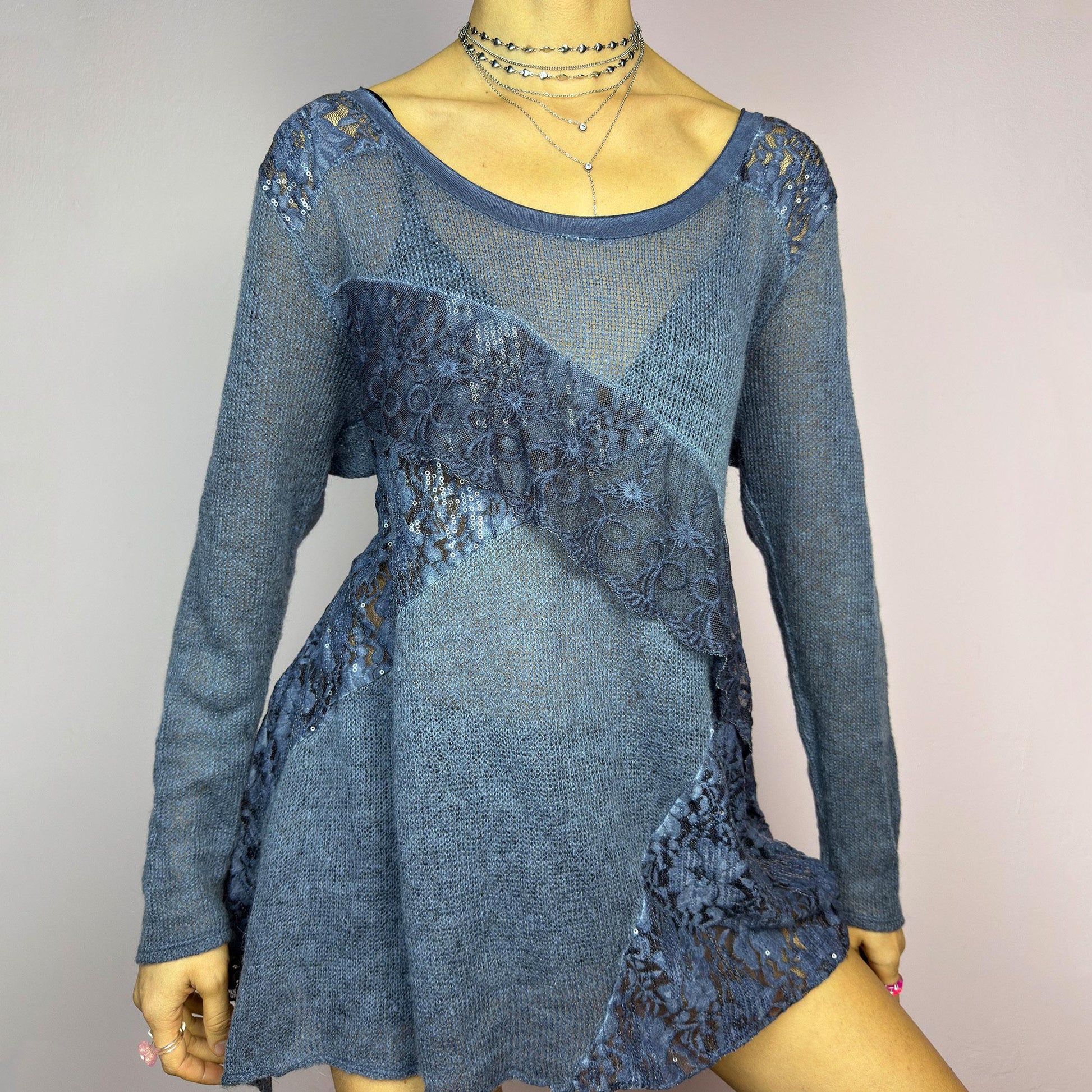 The Y2K Blue Lace Asymmetric Sweater is a vintage 2000s piece featuring a semi-sheer knit design with delicate lace and sequin paneling in an asymmetrical pattern. Designed with a scoop neckline, long sleeves, and a flowy silhouette, this sweater tunic adds a dreamy, romantic touch to any outfit. Perfect for layering in autumn and winter. adult world shop