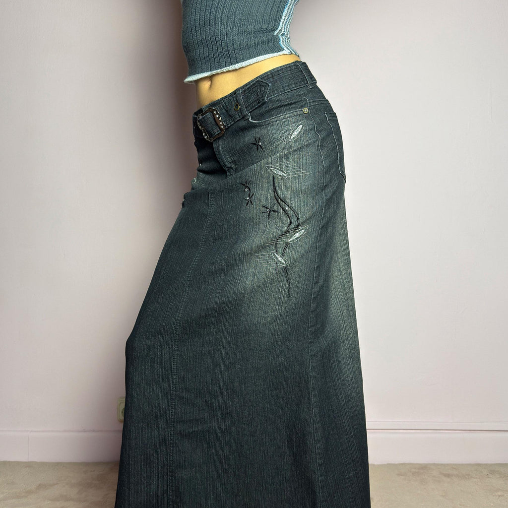 The Y2K Black Denim Maxi Skirt is a vintage 2000s piece featuring a sleek dark wash, rhinestone embellishments, and contrast stitching. Designed with a mid-rise fit and a stretchy denim fabric, it offers a structured silhouette that flares at the bottom. The skirt is finished with a button and zipper closure, pockets, and a removable belt for a customizable look. Perfect for casual streetwear outfits or an elegant night out. adult world shop