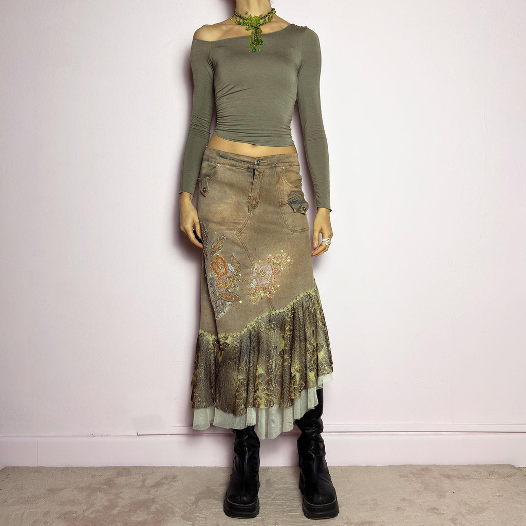 The Y2K Asymmetric Ruffle Midi Skirt is a vintage 2000s statement piece featuring a stretchy distressed tan denim base adorned with intricate beaded floral embroidery and sequin details. Designed with an asymmetric layered ruffle hem made of sheer lace and mesh with a metallic floral pattern, this skirt creates a dreamy, deconstructed aesthetic. It includes a button and zipper closure, belt loops, and pockets. adult world shop