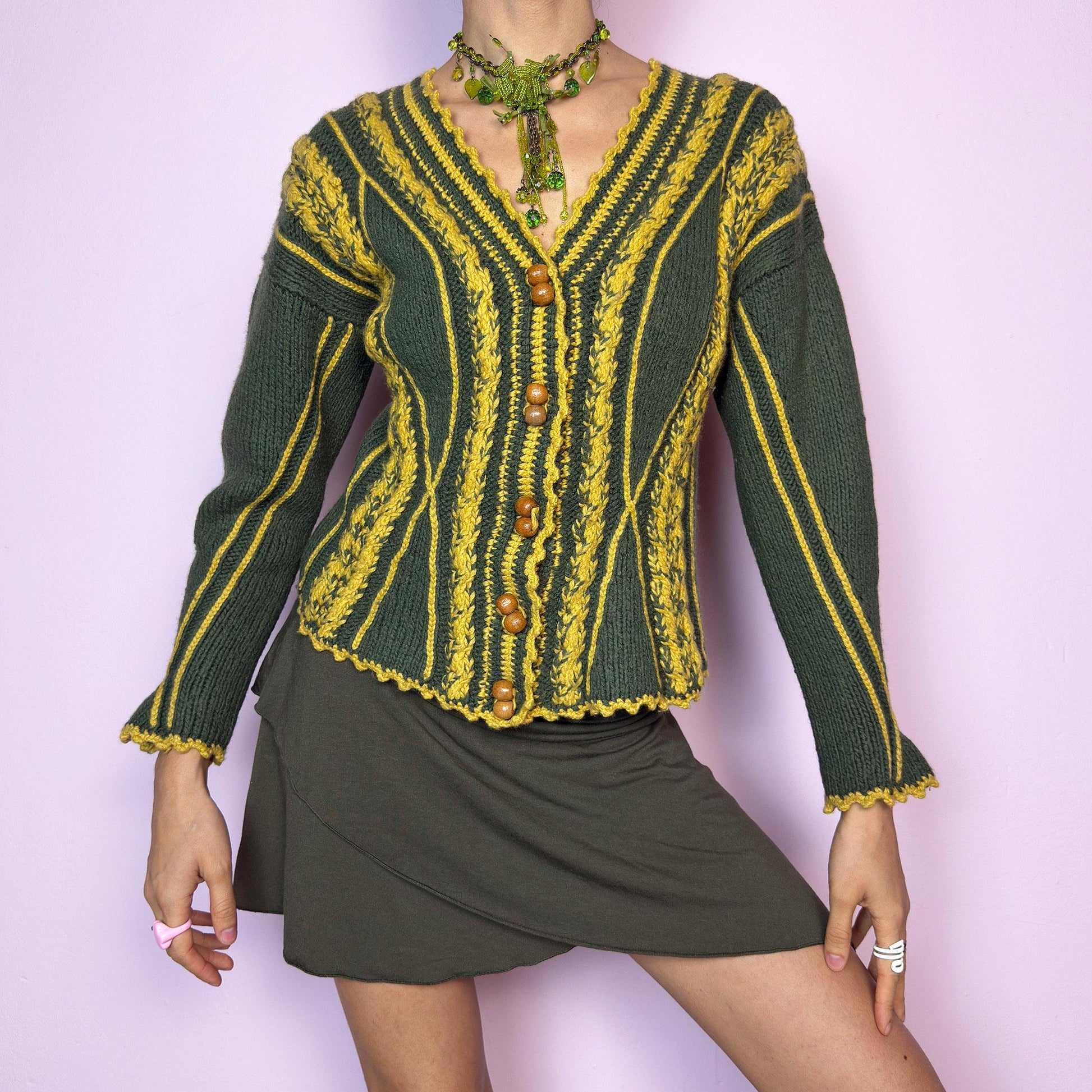 The Vintage Handmade-Style Chunky Knit Cardigan features intricate cable knit detailing in green and mustard yellow. With its flared cuffs, scalloped hems, and wooden buttons, this piece evokes a folk-inspired or alpine feel, reminiscent of 70s European ski towns and handmade market finds. The fitted silhouette and vertical lines create a flattering shape while adding bold texture. A one-of-a-kind layering piece perfect for forest walks, cozy days, or cottagecore styling.  adult world shop