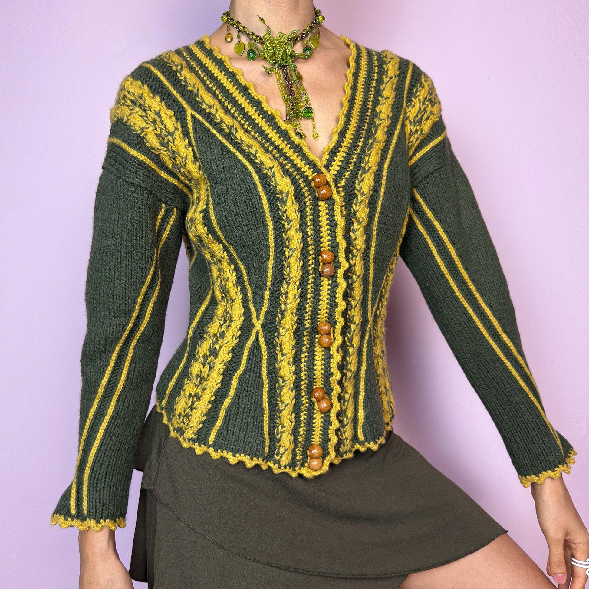 The Vintage Handmade-Style Chunky Knit Cardigan features intricate cable knit detailing in green and mustard yellow. With its flared cuffs, scalloped hems, and wooden buttons, this piece evokes a folk-inspired or alpine feel, reminiscent of 70s European ski towns and handmade market finds. The fitted silhouette and vertical lines create a flattering shape while adding bold texture. A one-of-a-kind layering piece perfect for forest walks, cozy days, or cottagecore styling.  adult world shop
