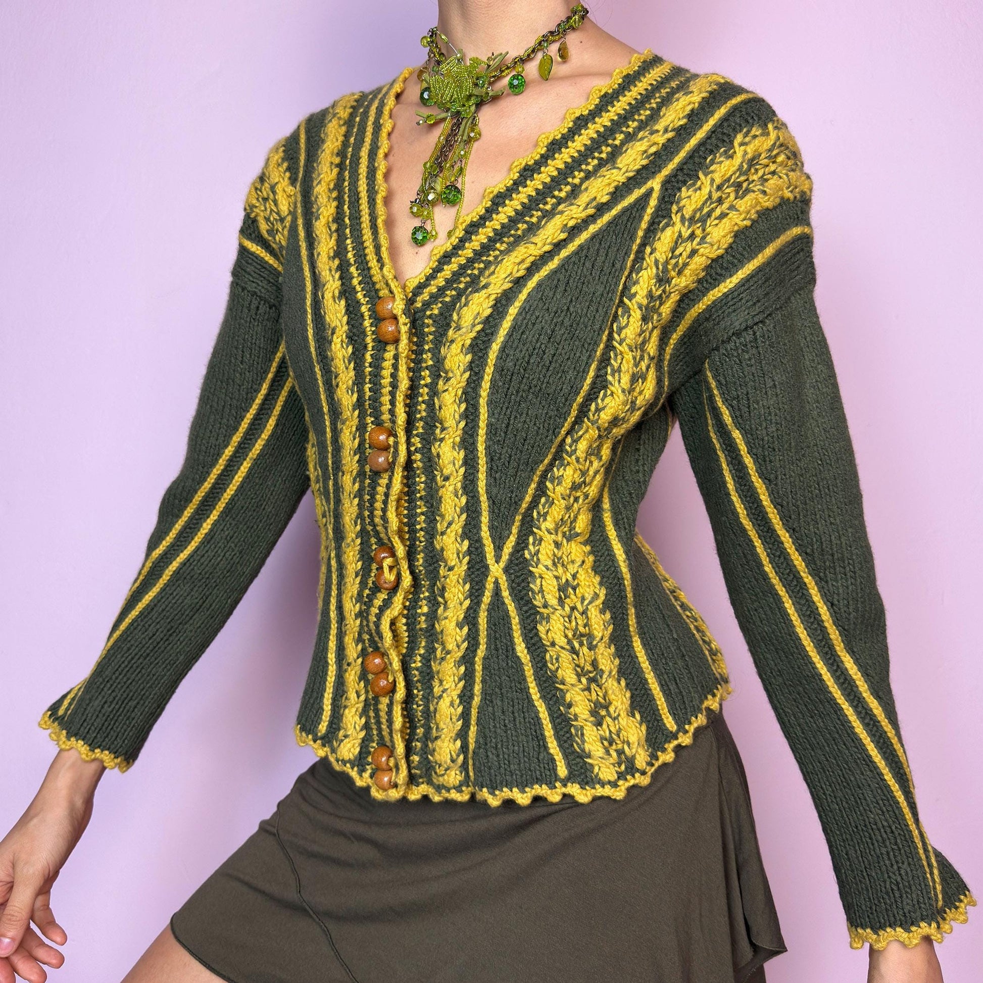 The Vintage Handmade-Style Chunky Knit Cardigan features intricate cable knit detailing in green and mustard yellow. With its flared cuffs, scalloped hems, and wooden buttons, this piece evokes a folk-inspired or alpine feel, reminiscent of 70s European ski towns and handmade market finds. The fitted silhouette and vertical lines create a flattering shape while adding bold texture. A one-of-a-kind layering piece perfect for forest walks, cozy days, or cottagecore styling.  adult world shop