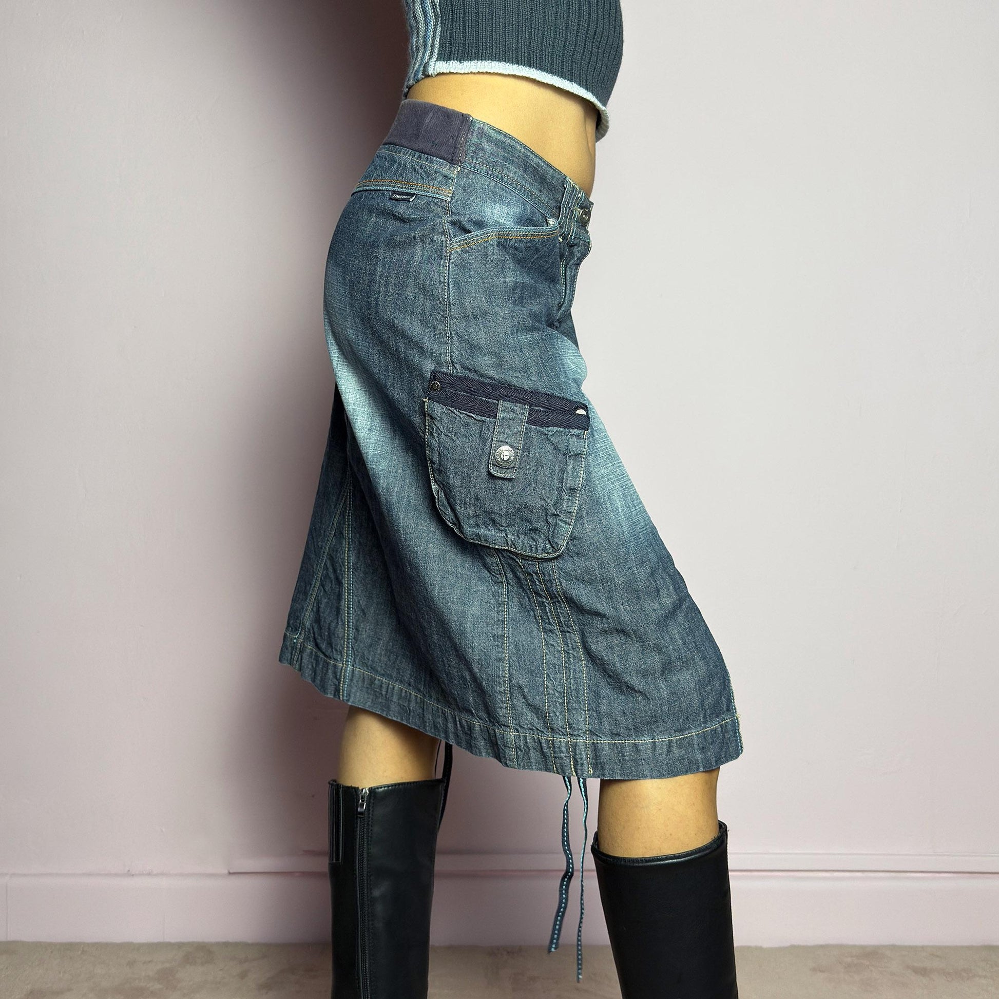 The Y2K Deconstructed Denim Midi Skirt is a vintage 2000s standout piece featuring a structured dark-wash denim fabric with contrast stitching. Designed with an asymmetrical front slit, utility-style cargo pockets, and adjustable drawstrings. Finished with a button and zipper closure, belt loops, and a mid-length silhouette, it&#39;s perfect for effortless grunge and utilitarian-inspired outfits. adult world shop