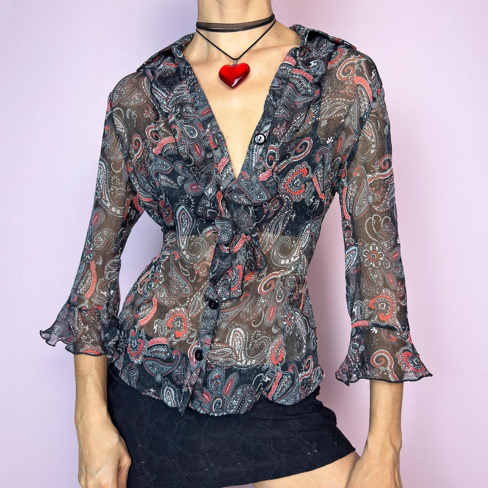 The Vintage 90s Black Ruffle Sheer Blouse features a semi-sheer fabric with an allover paisley print in red, grey, and white tones. This fitted silhouette includes three-quarter bell sleeves with flared ruffle cuffs, a romantic ruffled collar, and a front button-up closure. The lightweight fabric makes it perfect for summer layering or wearing on its own for a sleek, sexy vibe. Great vintage condition. adult world shop