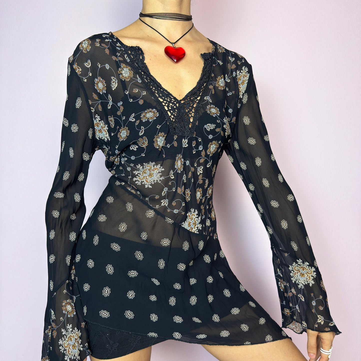 The Y2K Asymmetric Bell Sleeve Top is a vintage 2000s piece featuring a sheer black floral print with an asymmetric hem and dramatic bell sleeves for a flowy silhouette. The deep V-neckline is accented with crochet lace trim for a romantic touch. Lightweight and breathable, it’s perfect for summer layering or festival styling. Includes long sleeves with a bell flare for an elegant witchy vibe. Great vintage condition. adult world shop