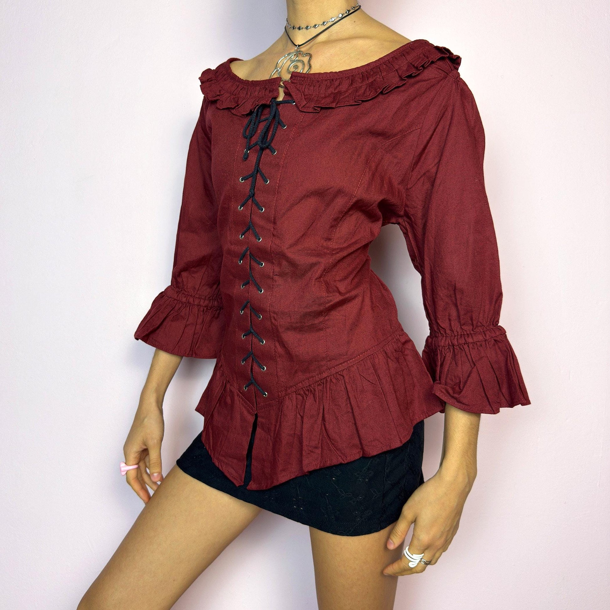 The Vintage 90s Maroon Lace Up Top is a romantic and dramatic piece inspired by gothic and cottagecore aesthetics. Crafted in a deep wine red tone, it features front and back corset-style lace-up details for an adjustable fit. The design includes a ruffled off-the-shoulder neckline, voluminous three-quarter sleeves with gathered ruffle cuffs, and a flattering cinched waist with a ruffled hem. Perfect for creating fairytale-inspired outfits or statement festival looks. adult world shop