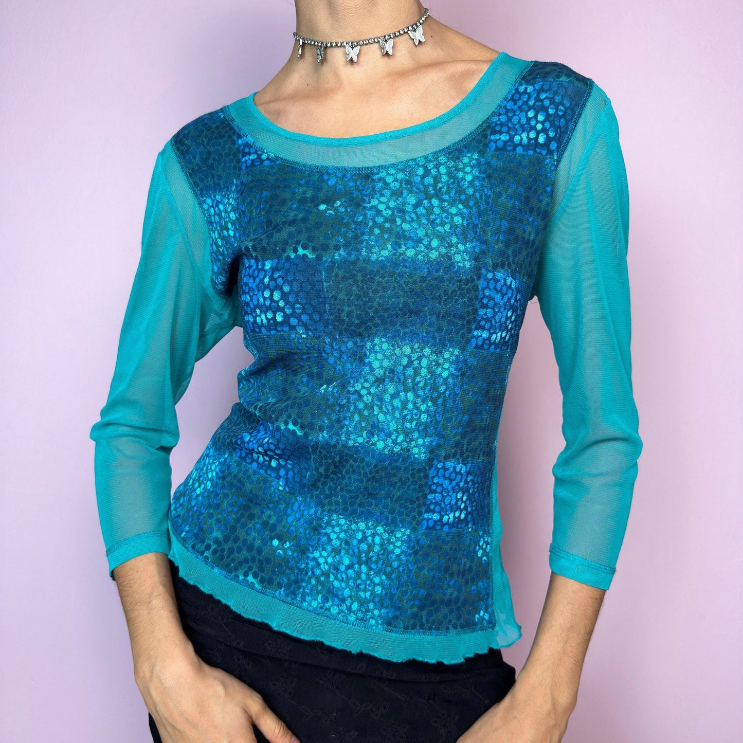 The Y2K Blue Patchwork Mesh Top is a vintage 2000s blue mesh top featuring a striking patchwork-style abstract print in shades of turquoise and electric blue. Designed with semi-sheer mesh sleeves and body, it has a fitted silhouette and three-quarter length sleeves. Lightweight and stretchy, this top is perfect for layering in rave, festival, or cyber looks. Excellent vintage condition.