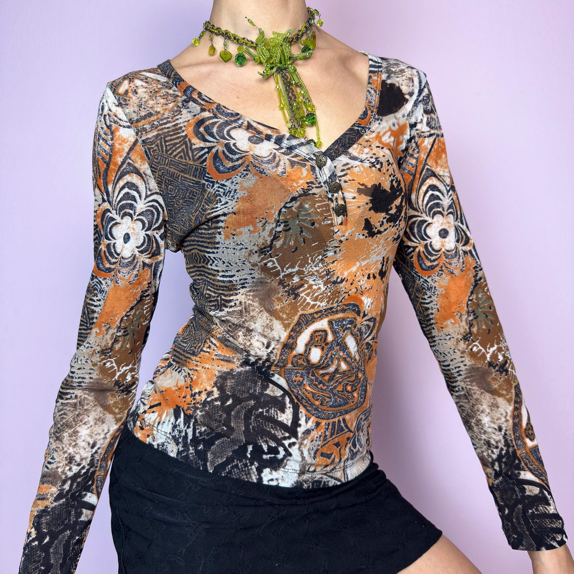 The Y2K Burnt Orange Mesh Top is a vintage 2000s long sleeve top featuring a bold tribal-inspired abstract print in a moody palette of burnt orange, black, and beige. Made from semi-sheer stretchy mesh, it hugs the body while staying breathable and comfy. Designed with a flattering V-neckline and three front buttons for added detail. Perfect for grunge, boho, or cybercore outfits – ideal as a statement piece or for layering. Excellent vintage condition.