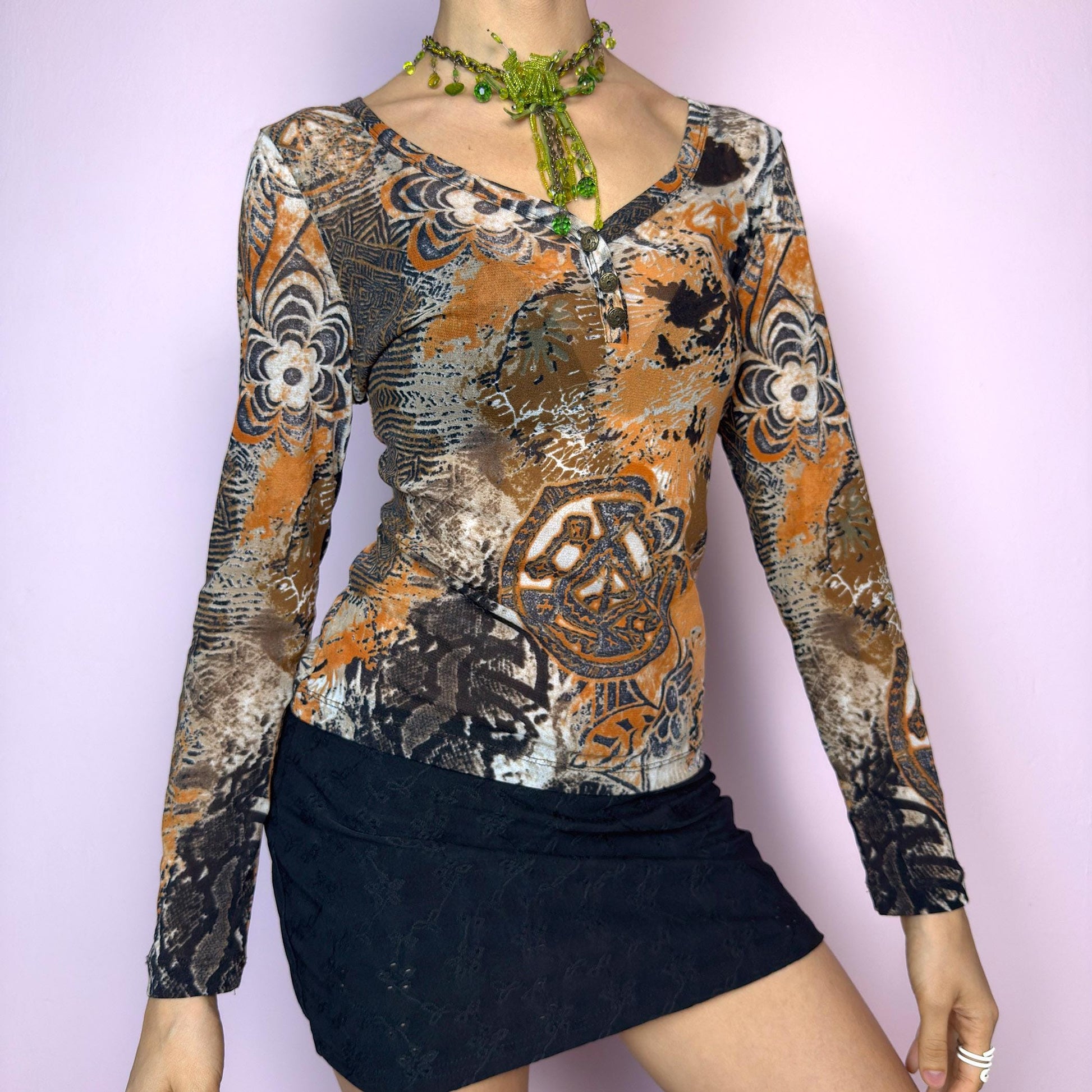 The Y2K Burnt Orange Mesh Top is a vintage 2000s long sleeve top featuring a bold tribal-inspired abstract print in a moody palette of burnt orange, black, and beige. Made from semi-sheer stretchy mesh, it hugs the body while staying breathable and comfy. Designed with a flattering V-neckline and three front buttons for added detail. Perfect for grunge, boho, or cybercore outfits – ideal as a statement piece or for layering. Excellent vintage condition.