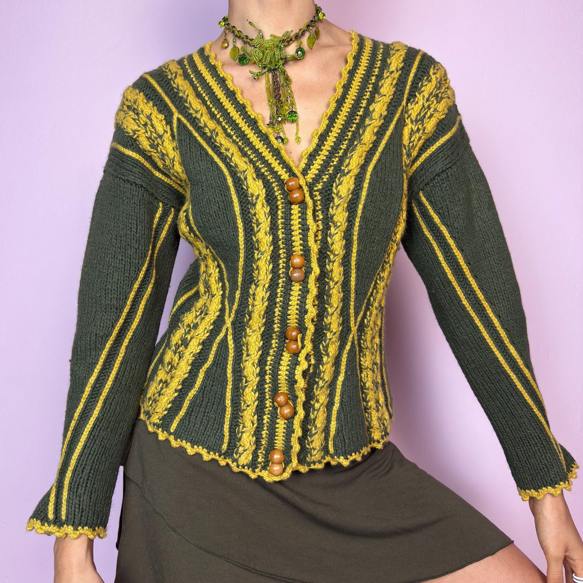 The Vintage Handmade-Style Chunky Knit Cardigan features intricate cable knit detailing in green and mustard yellow. With its flared cuffs, scalloped hems, and wooden buttons, this piece evokes a folk-inspired or alpine feel, reminiscent of 70s European ski towns and handmade market finds. The fitted silhouette and vertical lines create a flattering shape while adding bold texture. A one-of-a-kind layering piece perfect for forest walks, cozy days, or cottagecore styling.  adult world shop