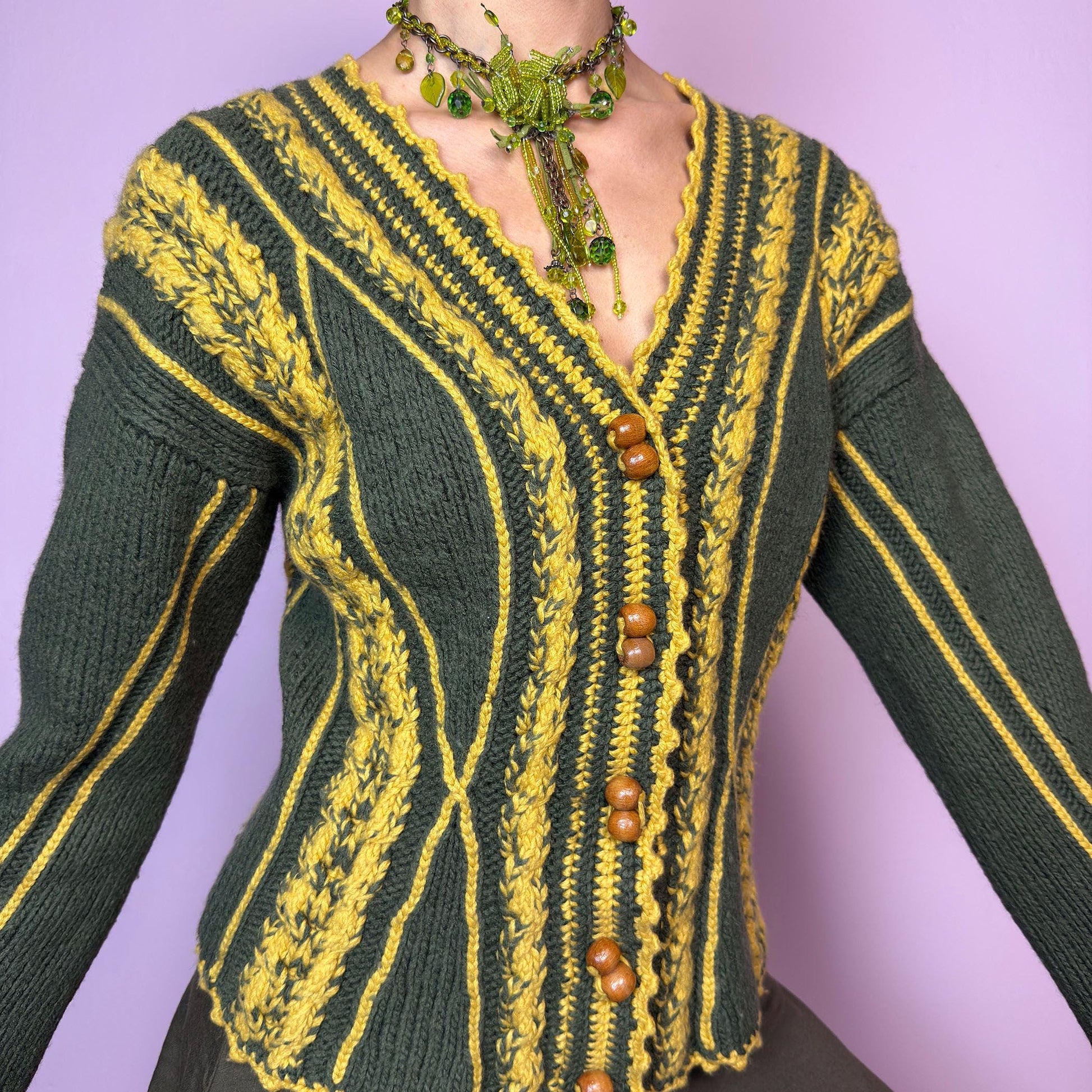 The Vintage Handmade-Style Chunky Knit Cardigan features intricate cable knit detailing in green and mustard yellow. With its flared cuffs, scalloped hems, and wooden buttons, this piece evokes a folk-inspired or alpine feel, reminiscent of 70s European ski towns and handmade market finds. The fitted silhouette and vertical lines create a flattering shape while adding bold texture. A one-of-a-kind layering piece perfect for forest walks, cozy days, or cottagecore styling.  adult world shop