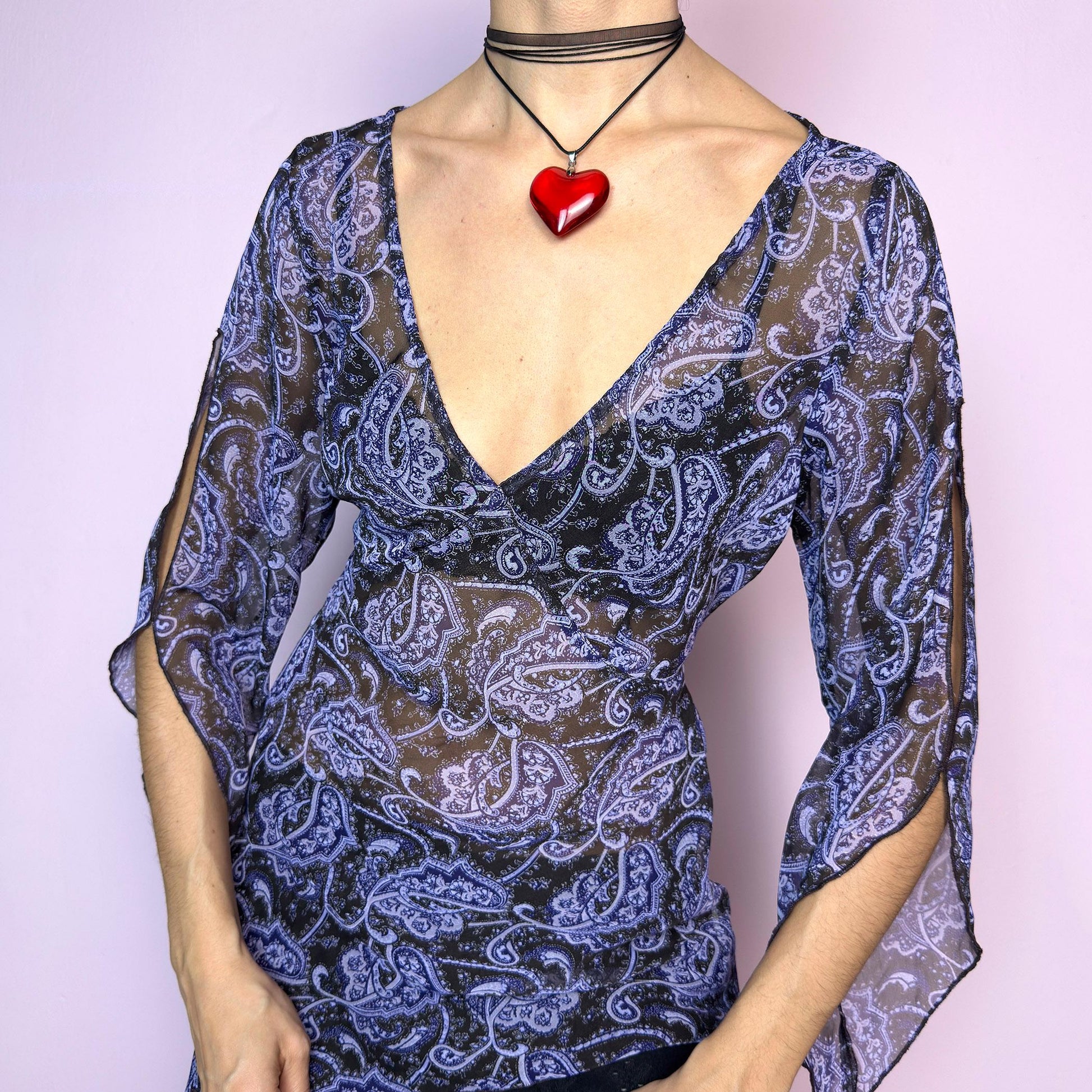 The Y2K Black Sheer Paisley Blouse is a vintage 2000s sheer paisley print blouse in black and purple tones. Features an asymmetrical hemline, open slit bell sleeves, and a flattering deep V-neckline with a tie-back detail to cinch the waist. Made from lightweight fabric for a breezy fit. Perfect for summer layering, night-out looks, or festival styling. Great vintage condition. adult world shop