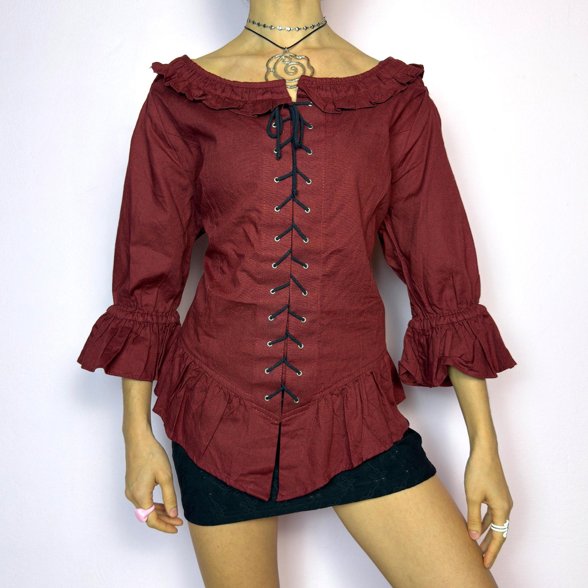 The Vintage 90s Maroon Lace Up Top is a romantic and dramatic piece inspired by gothic and cottagecore aesthetics. Crafted in a deep wine red tone, it features front and back corset-style lace-up details for an adjustable fit. The design includes a ruffled off-the-shoulder neckline, voluminous three-quarter sleeves with gathered ruffle cuffs, and a flattering cinched waist with a ruffled hem. Perfect for creating fairytale-inspired outfits or statement festival looks. adult world shop