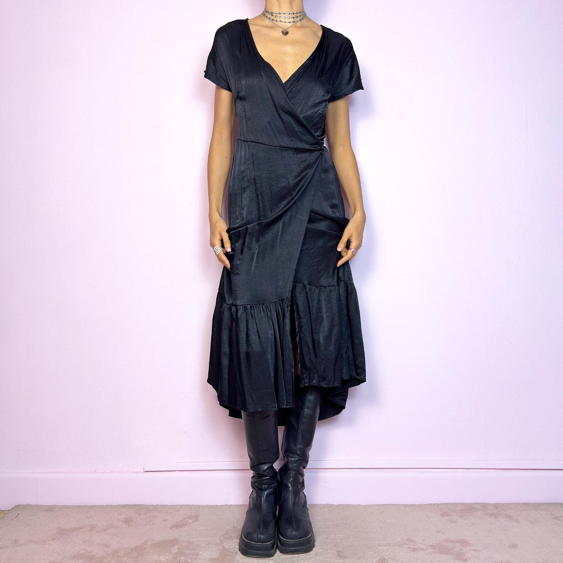 This vintage 2000s black wrap midi dress is made from a satin-style fabric with a soft sheen and airy drape. It features short sleeves, a plunging V-neckline, and an adjustable tie closure at the back. The hem is finished with a gathered ruffle panel, adding movement and shape. Perfect for elegant summer nights. A dramatic yet effortless piece with a quietly romantic silhouette.