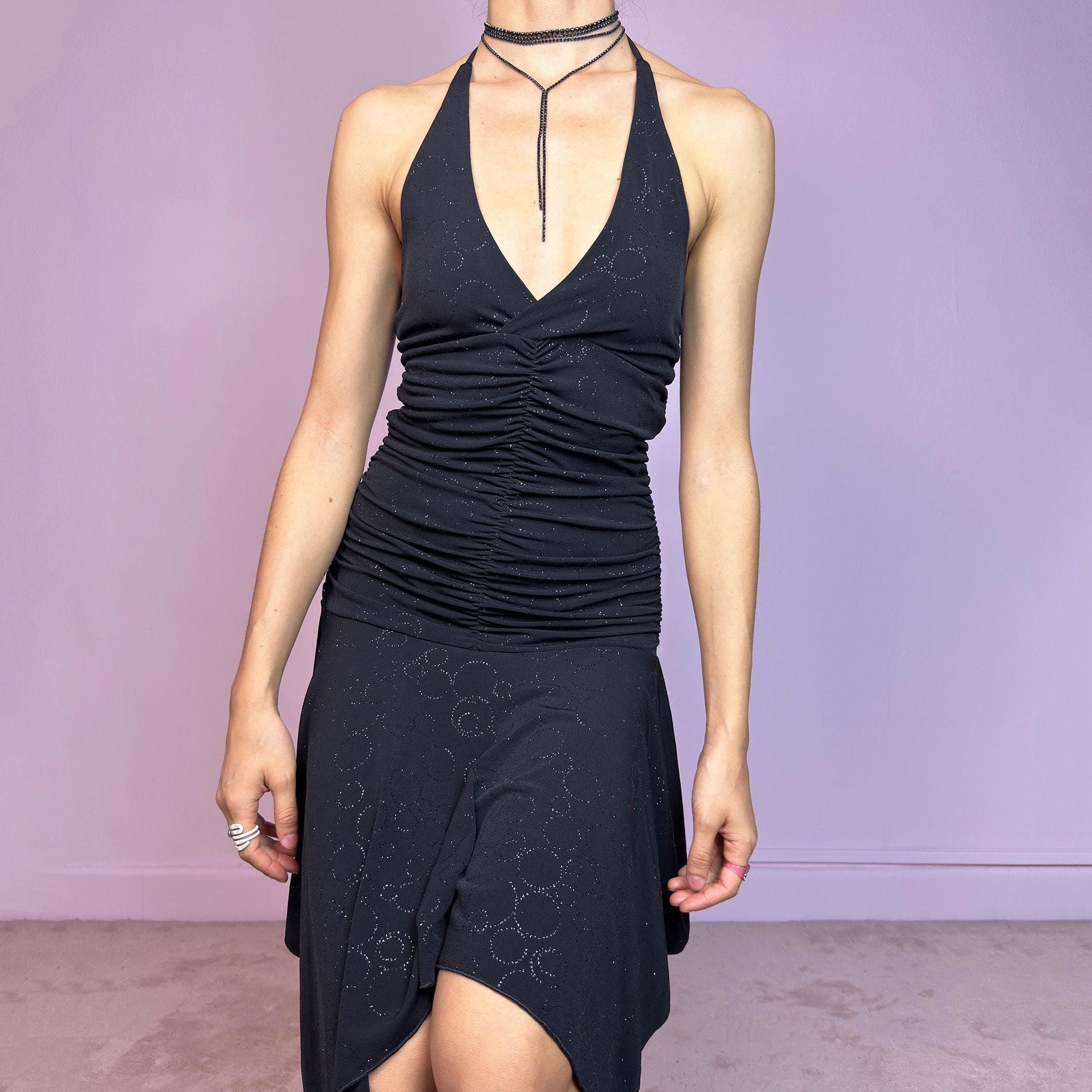 This vintage 2000s black dress features a halter tie neck, deep V-neckline, and allover subtle glitter swirl pattern. The ruched bodice sculpts the waist while the handkerchief hem adds movement and edge. Cut from stretchy fabric with a fluid, body-hugging drape. No closures. A cyber-glam silhouette with clubwear energy—perfect for parties, raves, or statement summer nights. Great vintage condition.