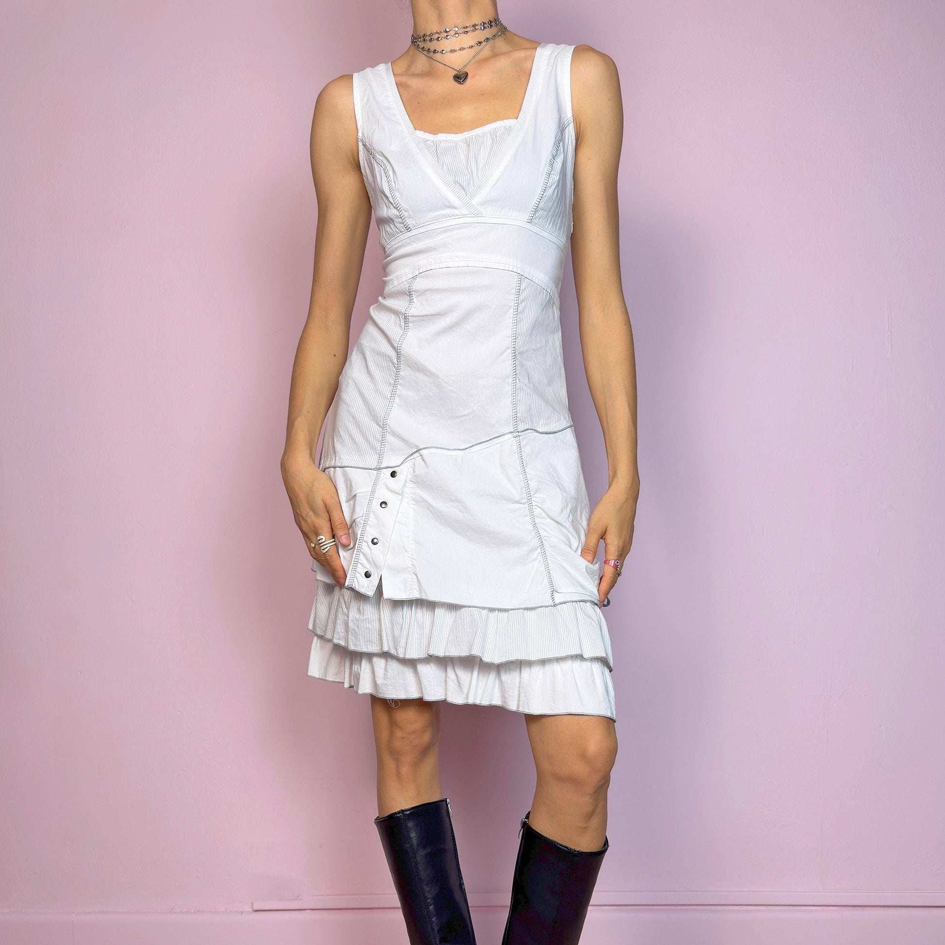 This vintage 2000s white sleeveless dress features a square neckline and sculpted panel seams. Made from a stretchy fabric, it includes a side zipper and falls to a midi length. Grommet hardware and a ruched drawstring side add a utilitarian twist, while the double-tiered ruffle hem softens the silhouette. Perfect for summer, this piece fuses deconstructed structure with a futuristic Y2K edge. Great vintage condition.