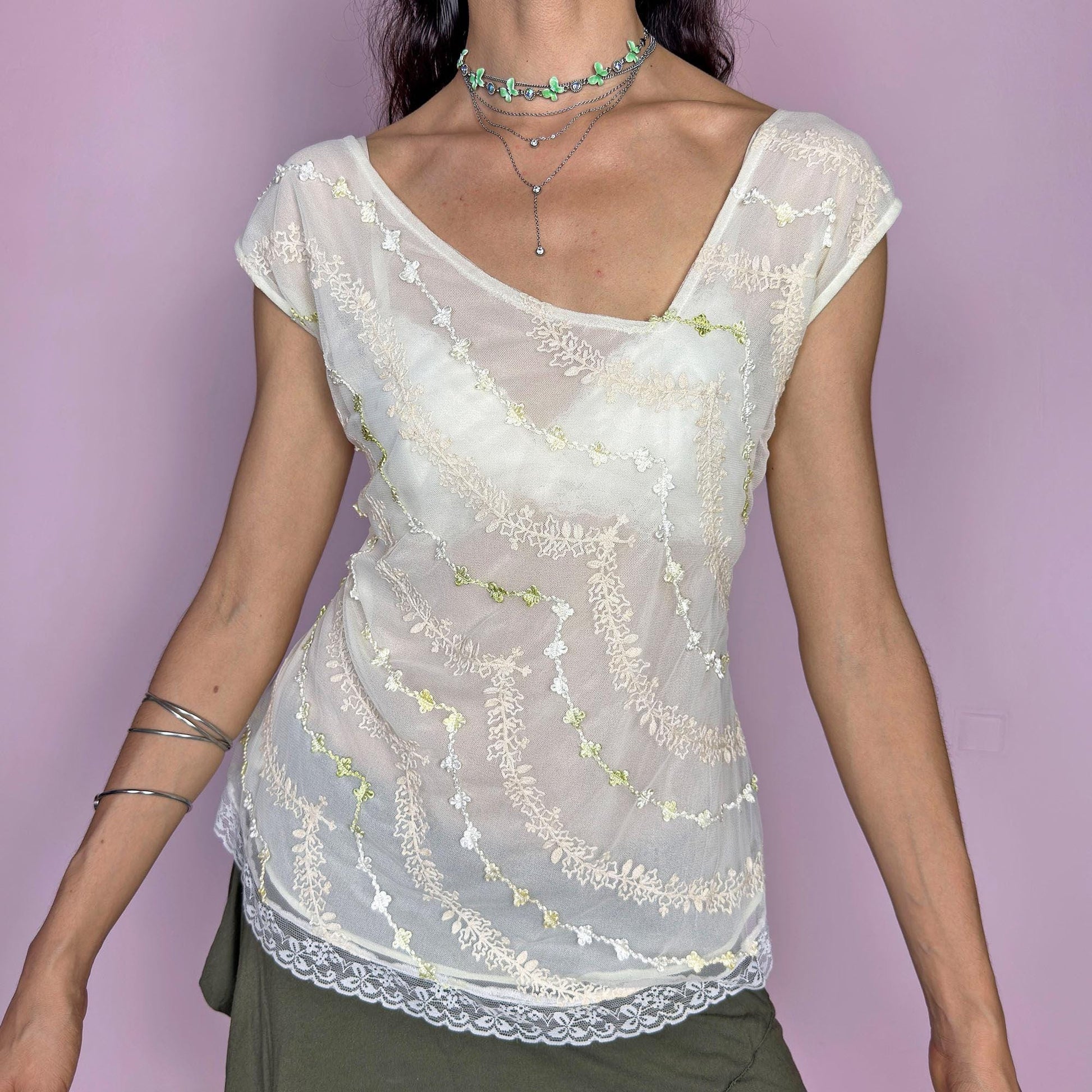 This vintage 2000s cream white mesh top features semi-sheer fabric with delicate embroidery in soft yellow, white, and pale green. The asymmetrical neckline and cap sleeves create an ethereal drape, while lace trim at the hem adds a romantic finish. This fairy-like Y2K blouse evokes lightness and whimsy—perfect for dreamy summer looks, warm days, and delicate layering. Made of polyester blend.