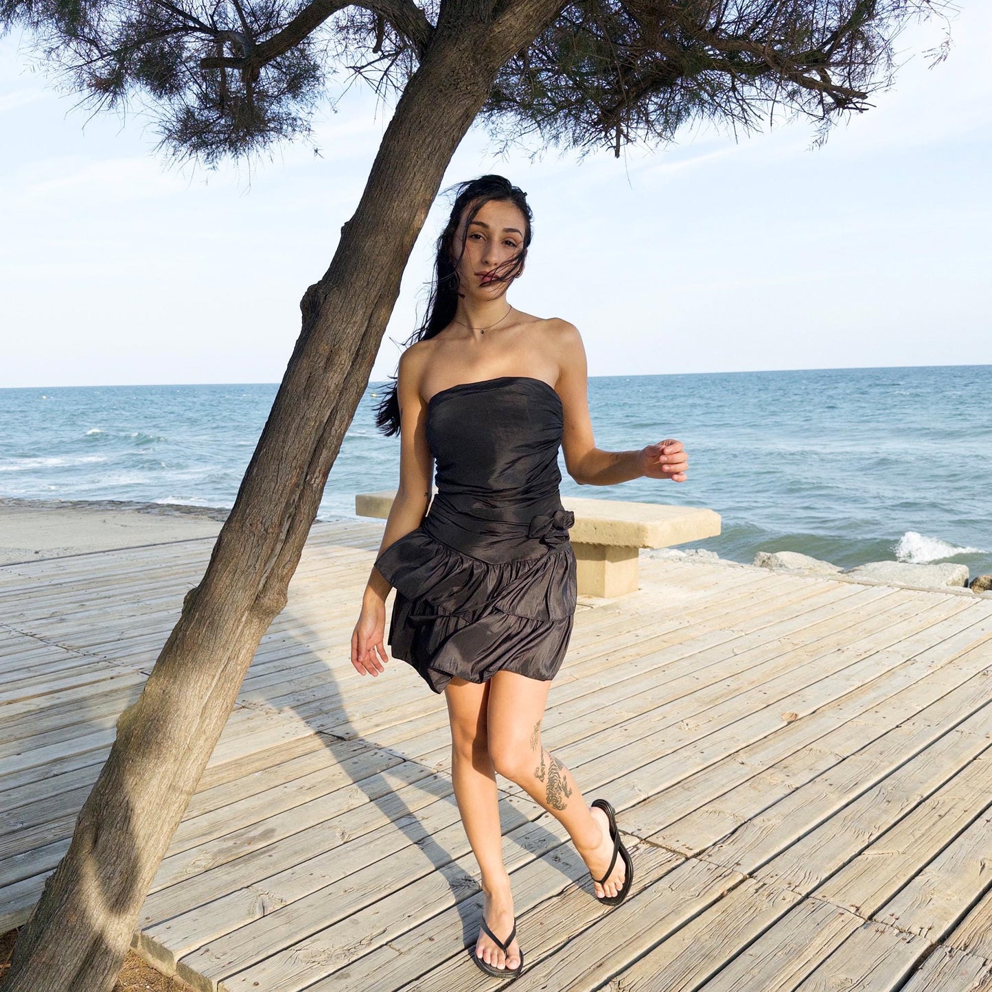 The vintage 90s black mini dress is a sculptural, strapless piece with ruched draping and voluminous double ruffle tiers. Made in Italy, it features a tie-back sash, shirred elastic panel, and side zip for a secure, flattering fit. Crafted from a polyester blend, it catches light effortlessly—ideal for summer nights, cocktail events, or rooftop parties. A super cute dress with high femme drama. Excellent vintage condition.