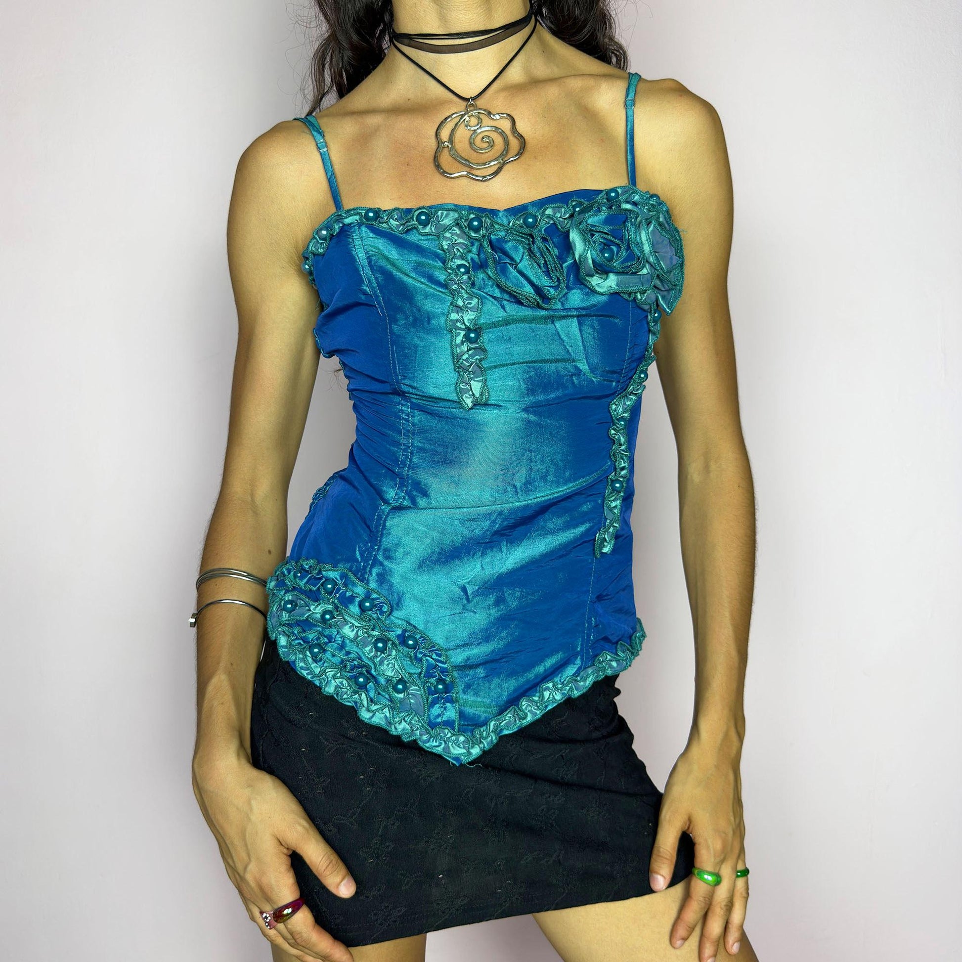 This vintage 2000s iridescent turquoise corset top features adjustable spaghetti straps, floral rosette appliqué, and beaded trim. The structured front sculpts the waist, while the ruched smocked back adds stretch and comfort. An asymmetrical hem and glossy finish elevate it for prom, weddings, or dreamy nights out. A romantic Y2K formal top that blends fairytale opulence with a bold, sculptural silhouette. Excellent vintage condition.