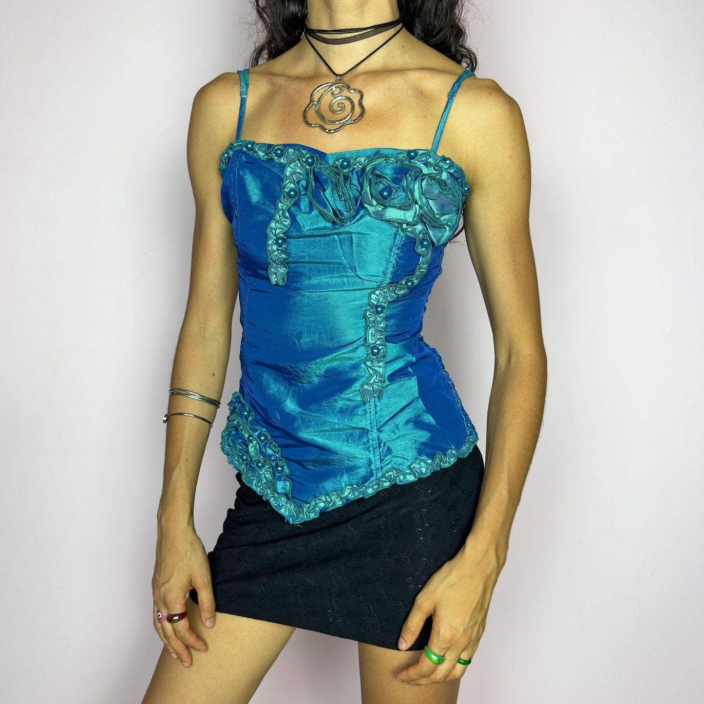 This vintage 2000s iridescent turquoise corset top features adjustable spaghetti straps, floral rosette appliqué, and beaded trim. The structured front sculpts the waist, while the ruched smocked back adds stretch and comfort. An asymmetrical hem and glossy finish elevate it for prom, weddings, or dreamy nights out. A romantic Y2K formal top that blends fairytale opulence with a bold, sculptural silhouette. Excellent vintage condition.