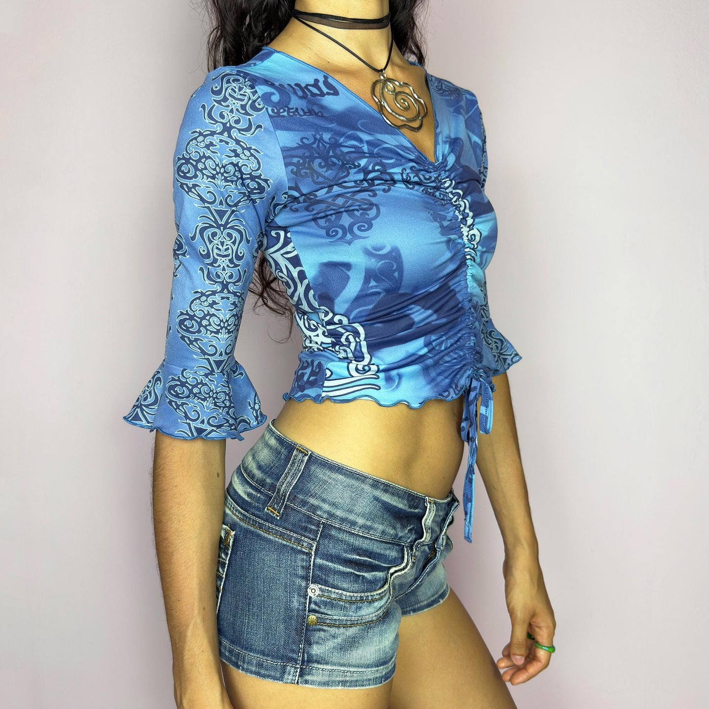 This vintage 2000s crop top features a bold tribal-inspired blue print with ruched detailing and an adjustable drawstring tie front. Designed with three-quarter bell sleeves and a fitted silhouette, it captures cyber Y2K energy perfect for festivals, parties, and summer styling. A rare statement piece blending unique printwork with a flattering cut. Made from stretch fabric. Great vintage condition.