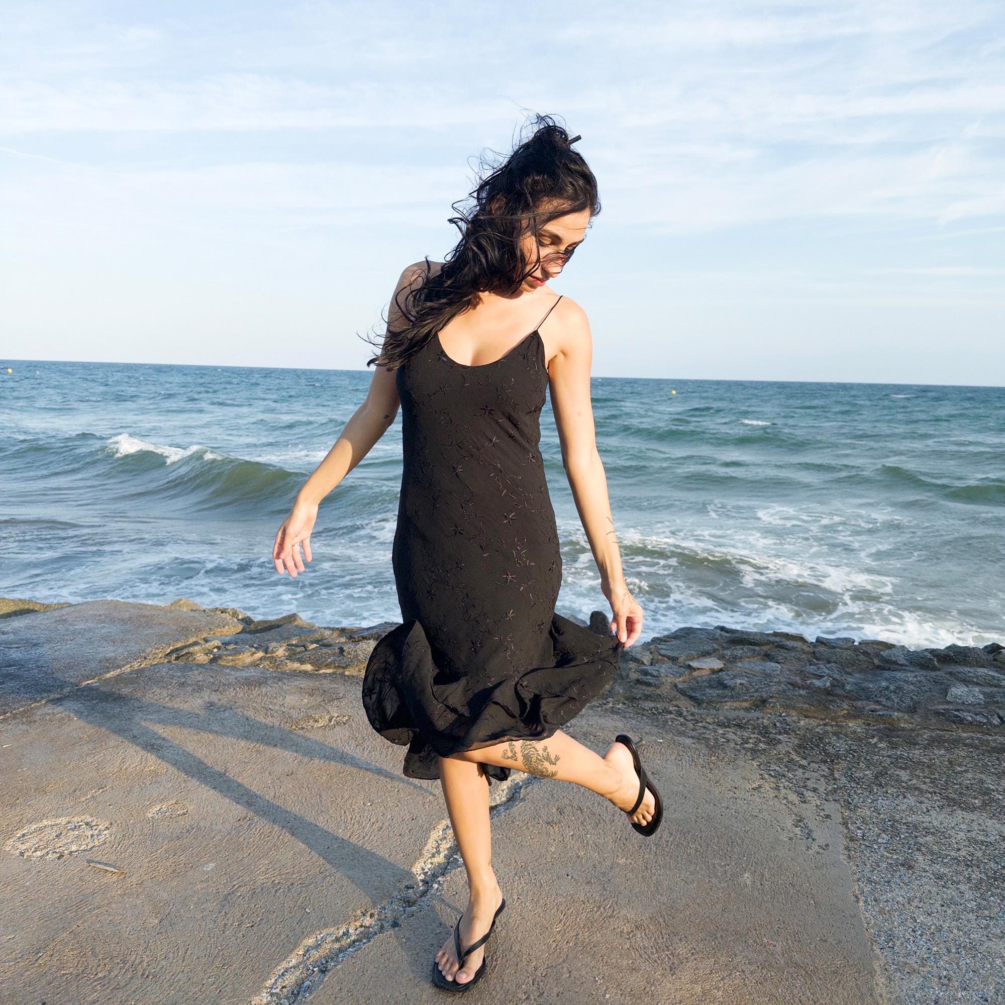 The vintage 2000s black midi dress is a French-made slip silhouette with delicate spaghetti straps and a tiered ruffle hem that drapes asymmetrically. Lightweight with subtle floral embroidery, it flows with ease—ideal for summer promenades, cocktail sunsets, or barefoot evenings. Its understated sensuality and effortless cut evoke a romantic calm. Excellent vintage condition.