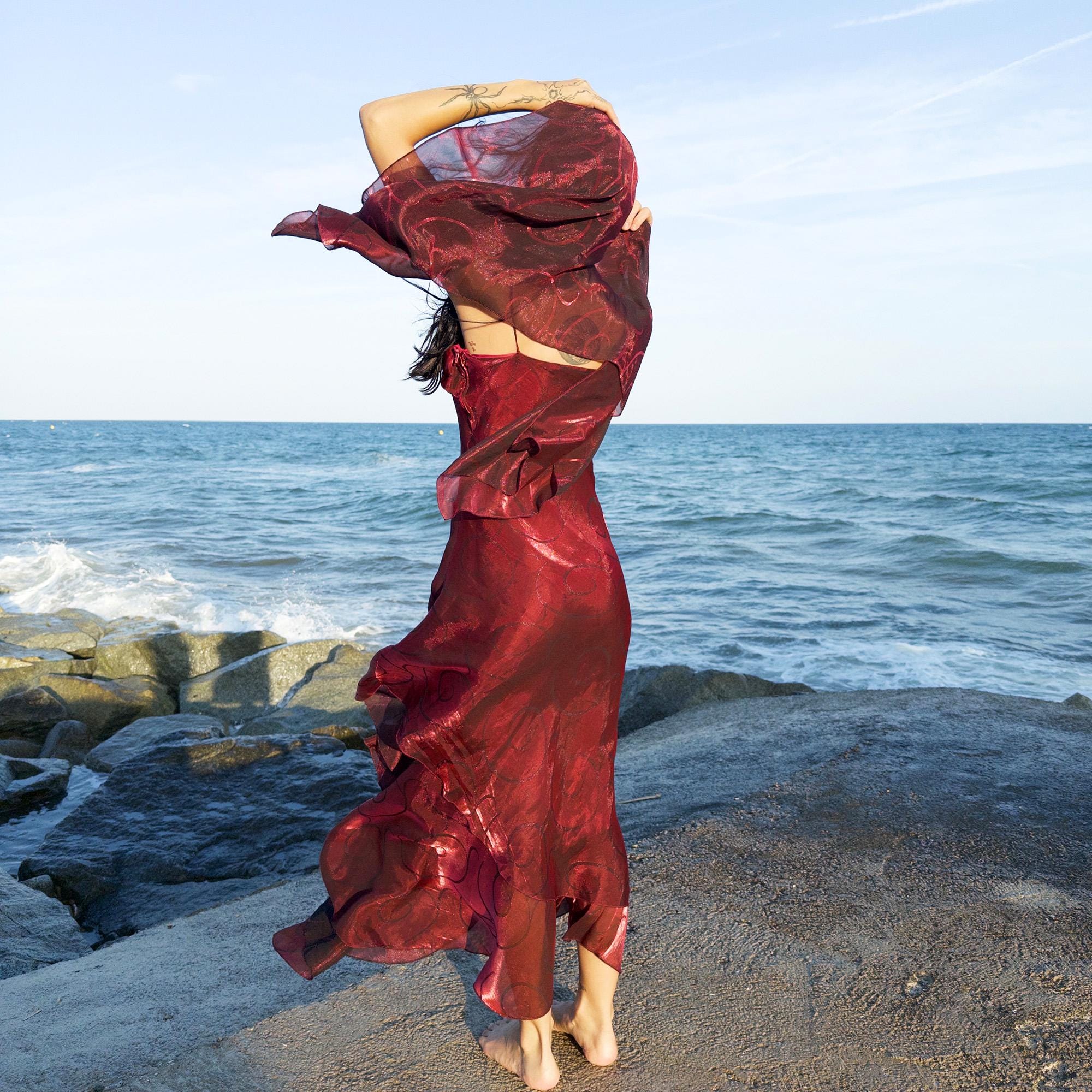 The vintage 2000s red organza-style slip midi dress is a French-made evening piece with layered maroon tones and tonal swirl pattern. With delicate spaghetti straps, side zip, and a flowing asymmetrical hem, it includes a matching scarf. Ideal for galas, proms, or romantic nights under open skies. Crafted from a semi-sheer, non-stretch fabric. Excellent vintage condition.