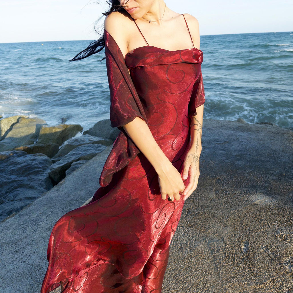 The vintage 2000s red organza-style slip midi dress is a French-made evening piece with layered maroon tones and tonal swirl pattern. With delicate spaghetti straps, side zip, and a flowing asymmetrical hem, it includes a matching scarf. Ideal for galas, proms, or romantic nights under open skies. Crafted from a semi-sheer, non-stretch fabric. Excellent vintage condition.