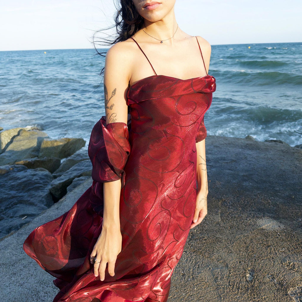 The vintage 2000s red organza-style slip midi dress is a French-made evening piece with layered maroon tones and tonal swirl pattern. With delicate spaghetti straps, side zip, and a flowing asymmetrical hem, it includes a matching scarf. Ideal for galas, proms, or romantic nights under open skies. Crafted from a semi-sheer, non-stretch fabric. Excellent vintage condition.