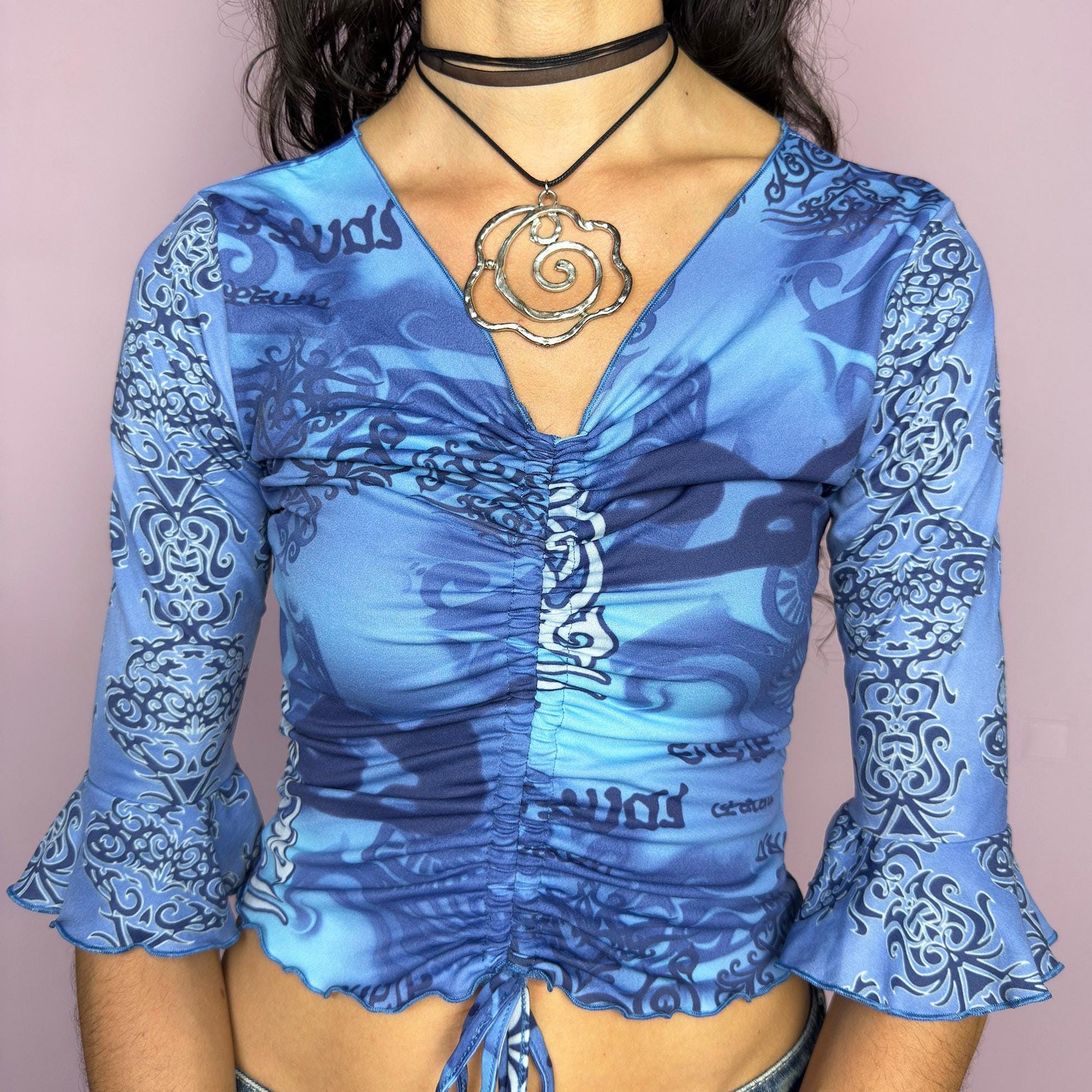 This vintage 2000s crop top features a bold tribal-inspired blue print with ruched detailing and an adjustable drawstring tie front. Designed with three-quarter bell sleeves and a fitted silhouette, it captures cyber Y2K energy perfect for festivals, parties, and summer styling. A rare statement piece blending unique printwork with a flattering cut. Made from stretch fabric. Great vintage condition.