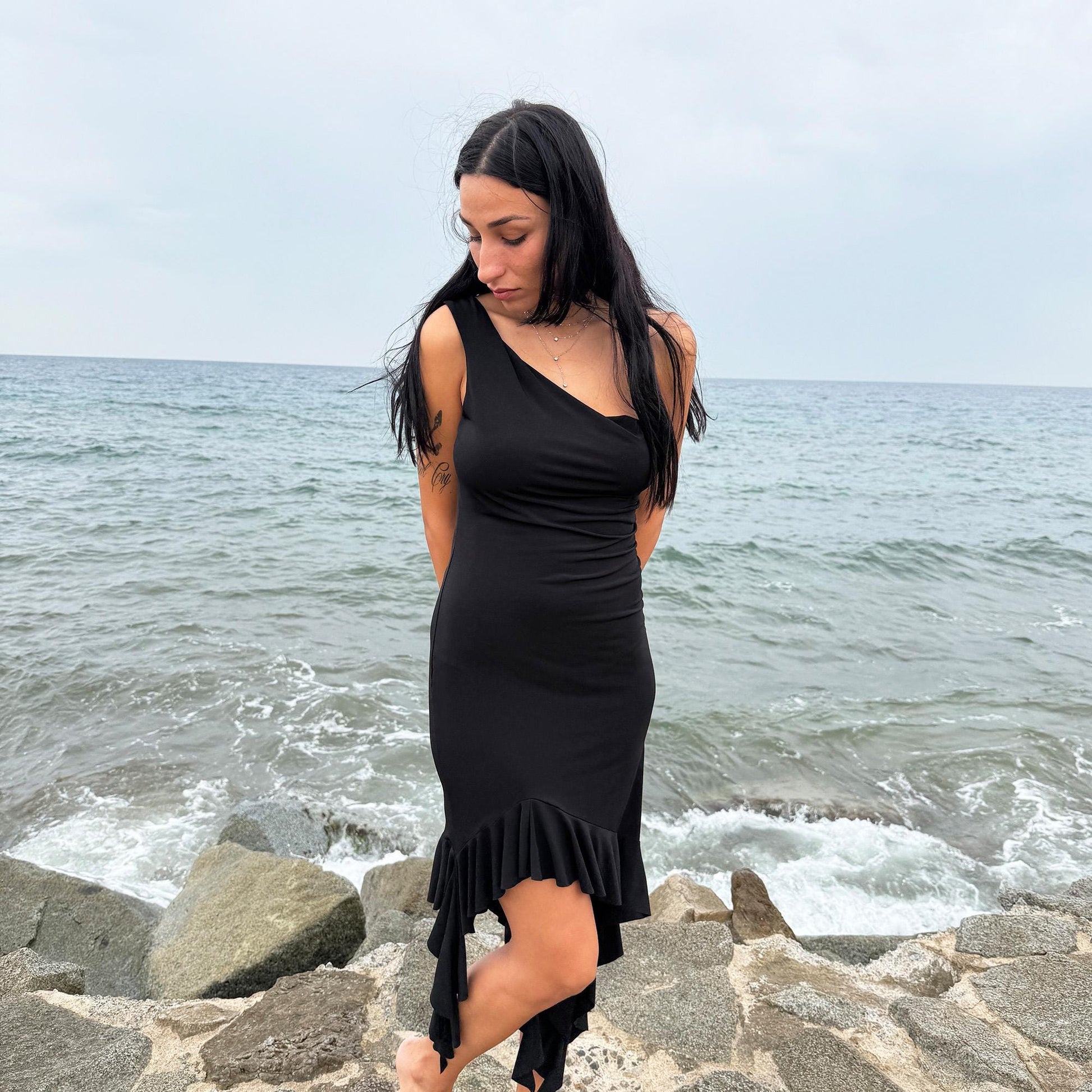 This vintage 2000s black mini dress features asymmetric straps with a subtle cowl neckline and a dramatic ruffled hem. Crafted from a stretchy fabric, it flatters the body with a bold party silhouette. Perfect for summer nights, club looks, or diva-inspired Y2K styling, this piece captures iconic 2000s glamour. Excellent vintage condition.