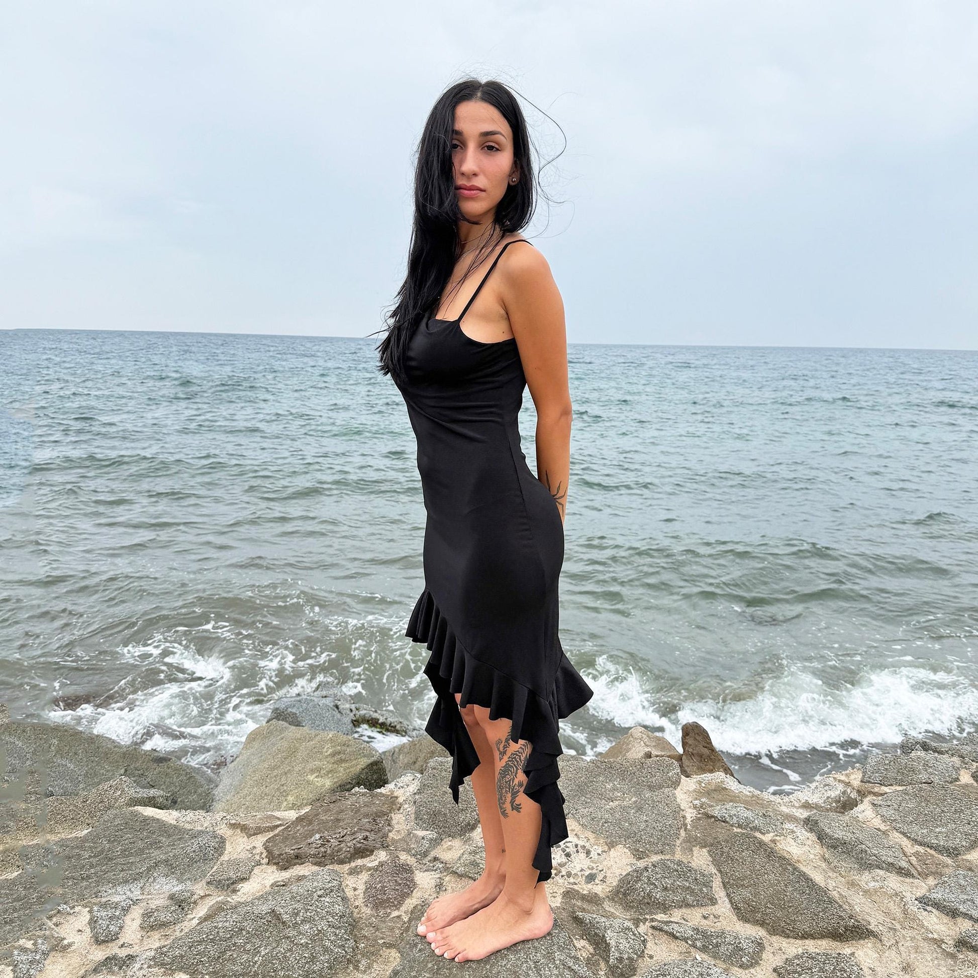 This vintage 2000s black mini dress features asymmetric straps with a subtle cowl neckline and a dramatic ruffled hem. Crafted from a stretchy fabric, it flatters the body with a bold party silhouette. Perfect for summer nights, club looks, or diva-inspired Y2K styling, this piece captures iconic 2000s glamour. Excellent vintage condition.